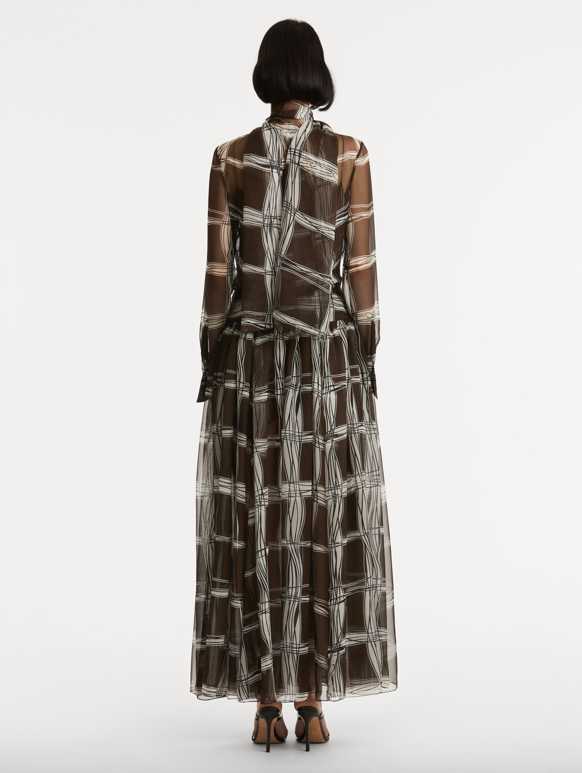 Full body back view of silk chiffon dress which features a brown base with a hand-drawn plaid pattern. Dress has long sleeves and a mock neckline with a self-tie closure. 