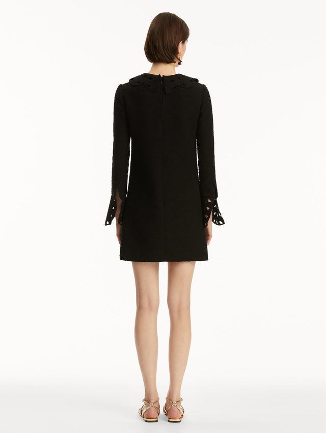 Model wearing black tweed long sleeve mini dress with threadwork cutouts at the scalloped collar and cuffs. Back view