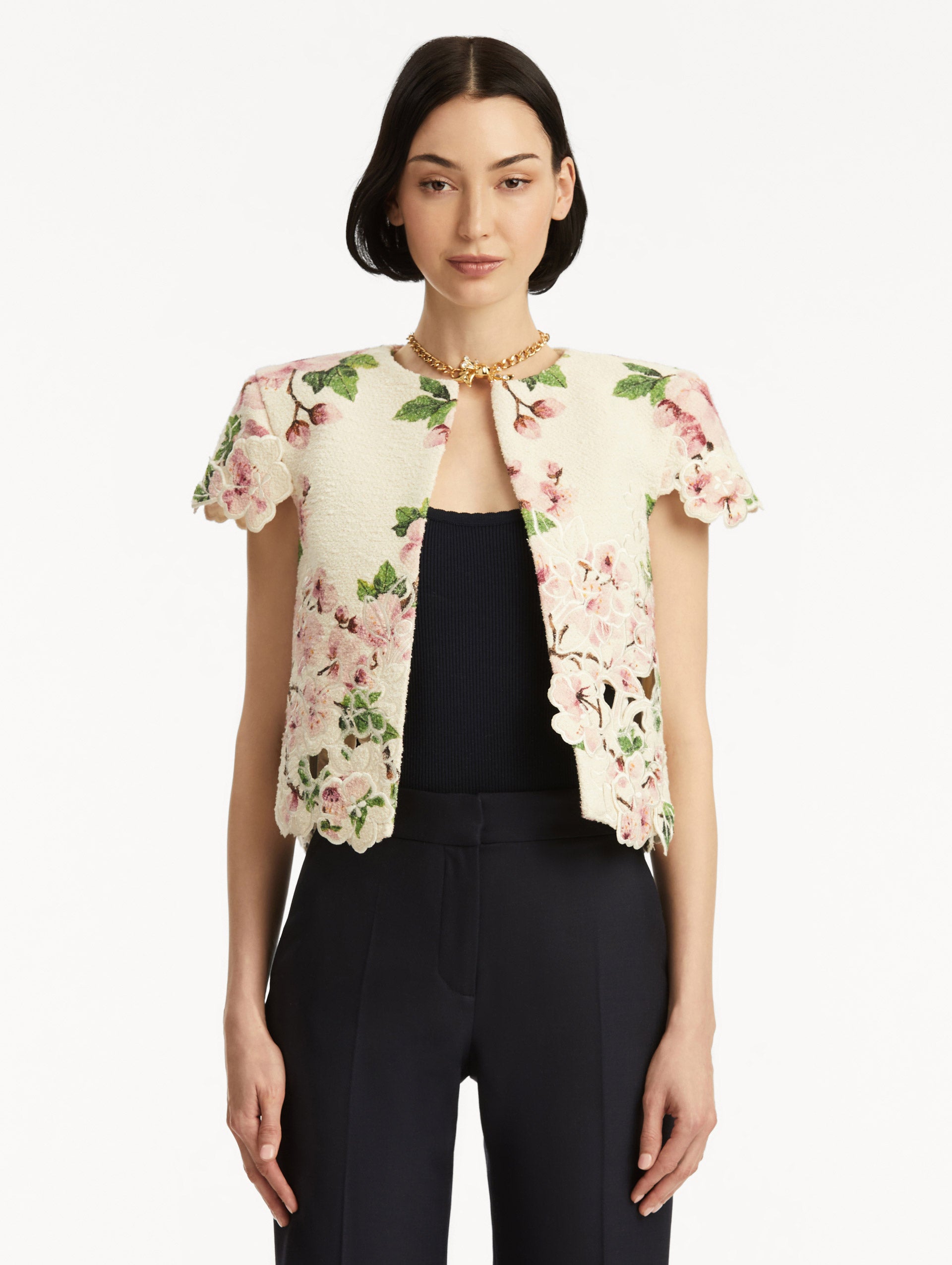 Model wearing white short sleeve tweed jacket with pink cherry blossoms embroidery. Paired with black pants and tank. Front view