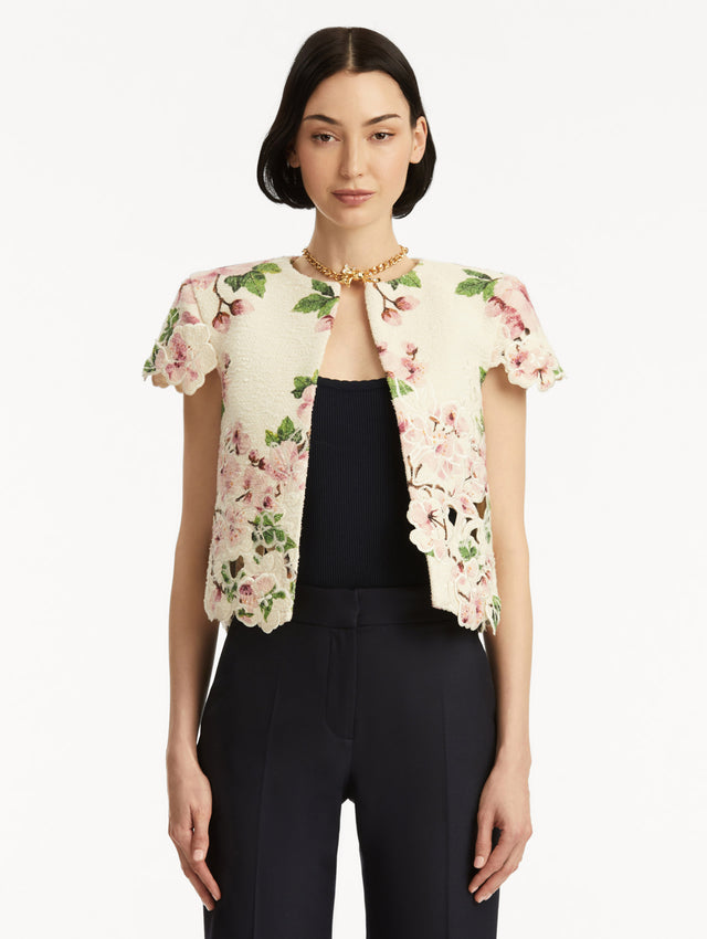 Model wearing white short sleeve tweed jacket with pink cherry blossoms embroidery. Paired with black pants and tank. Front view