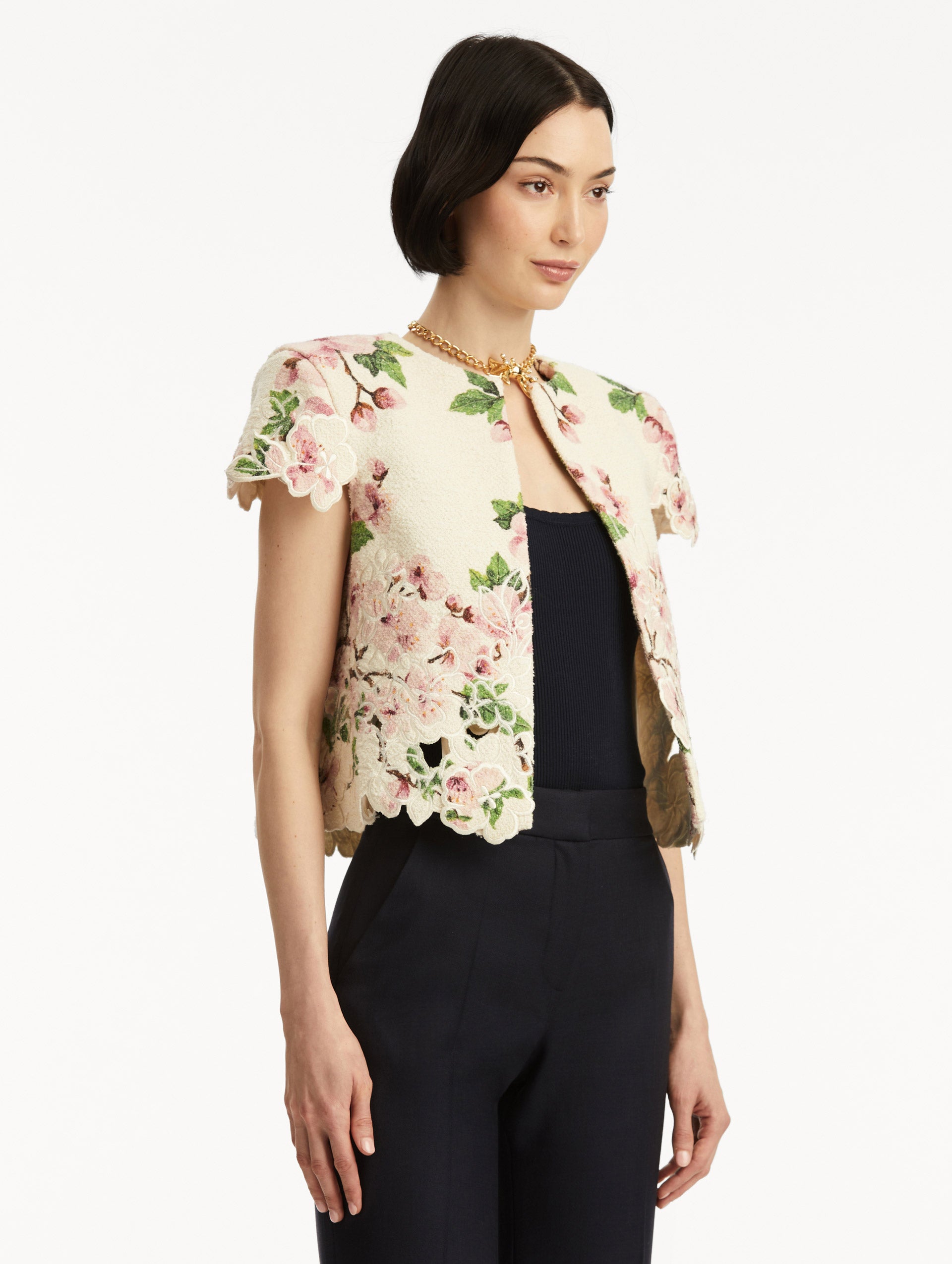 Model wearing white short sleeve tweed jacket with pink cherry blossoms embroidery. Paired with black pants and tank. Side view