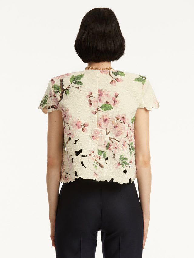 Model wearing white short sleeve tweed jacket with pink cherry blossoms embroidery. Paired with black pants and tank. Back view