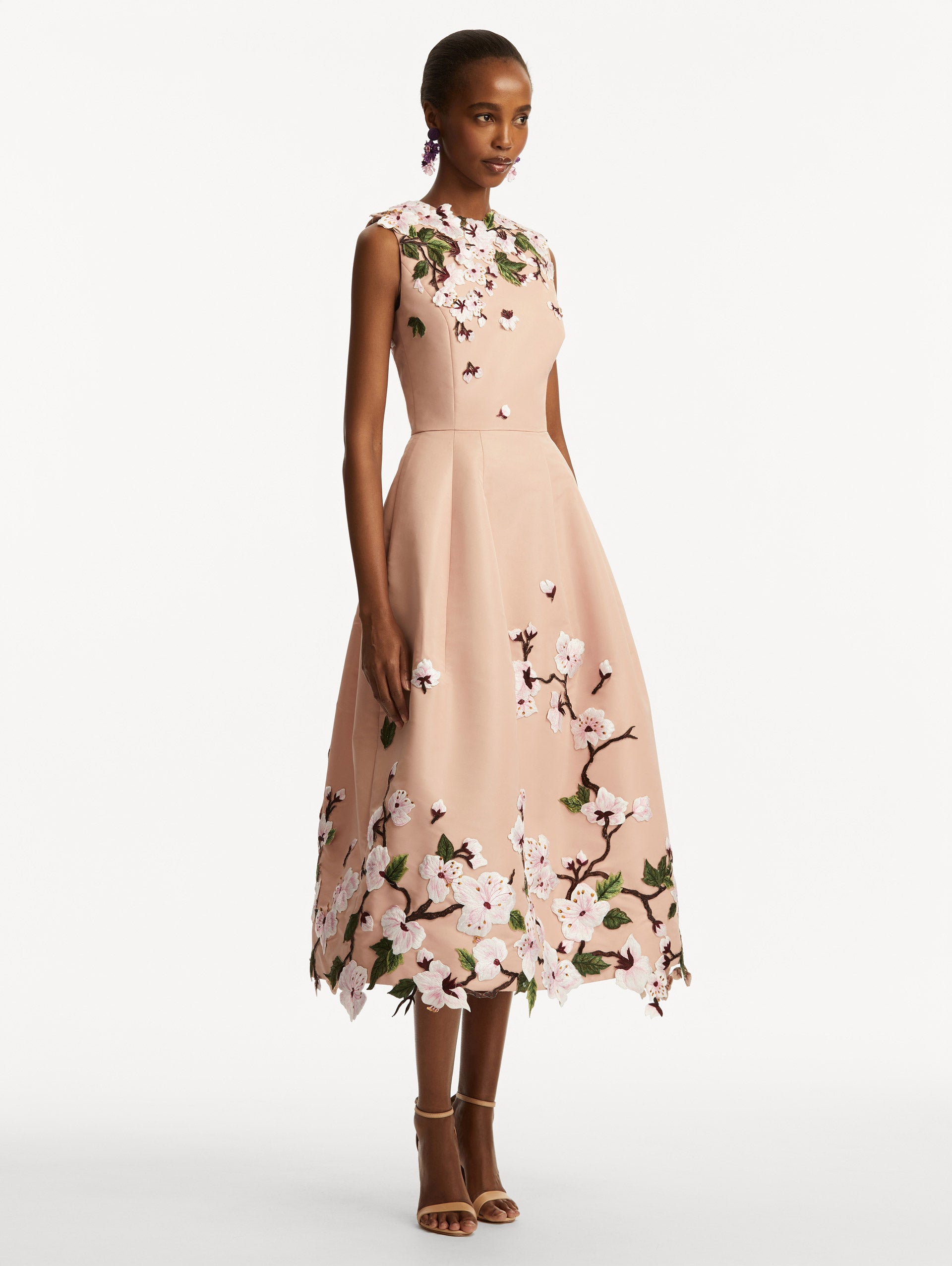 Model wearing a blush pink sleeveless dress with cherry blossoms embroidered throughout the top and bottom. Side view
