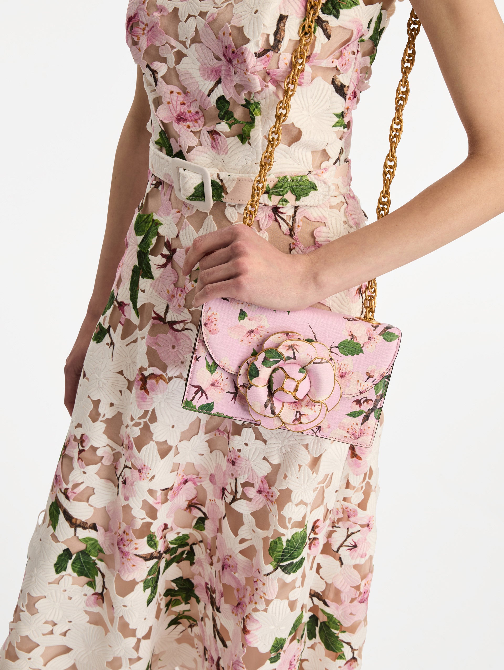The model is holding the TRO handbag in pink. This bag has a cherry blossom print all over and has a three-dimensional flower at the center clasp. The bag comes with a gold chain as a strap, and it is paired with a grey cherry blossom cardigan. Front View