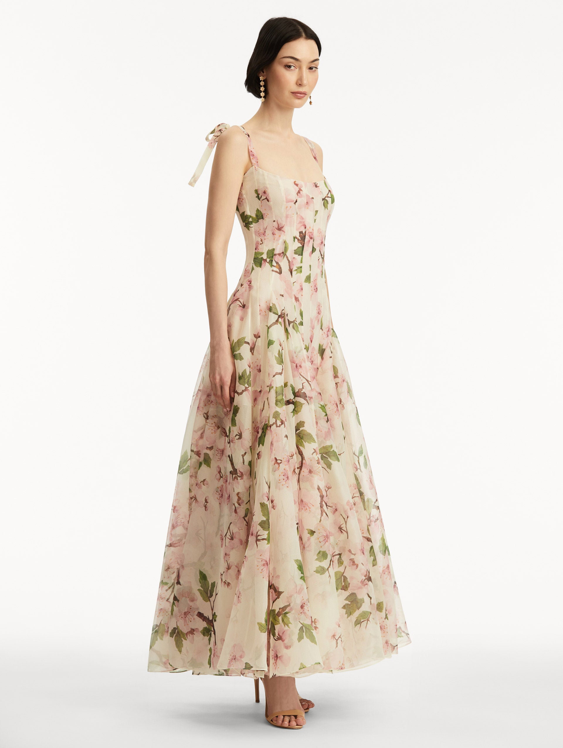 Model wearing White sleeveless gown with pink cherry blossoms, and bow detail in the straps. Side view.