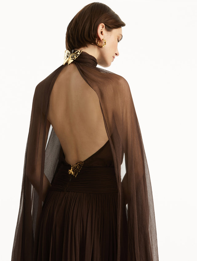 Brown chiffon gown with halter neck and long cascading sheer sleeves down the floor. The gown has an open back and a gold butterfly adornment at the closure around the neck and in the bottom of the back. Back view