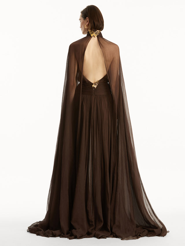 Brown chiffon gown with halter neck and long cascading sheer sleeves down the floor. The gown has an open back and a gold butterfly adornment at the closure around the neck and in the bottom of the back. Back view