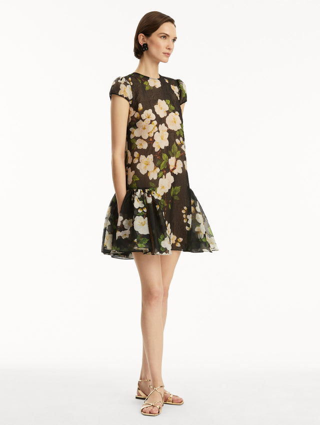 The model is wearing a short sleeve mini drop-waist dress in sheer black with white cherry blossoms printed all over. Side view.