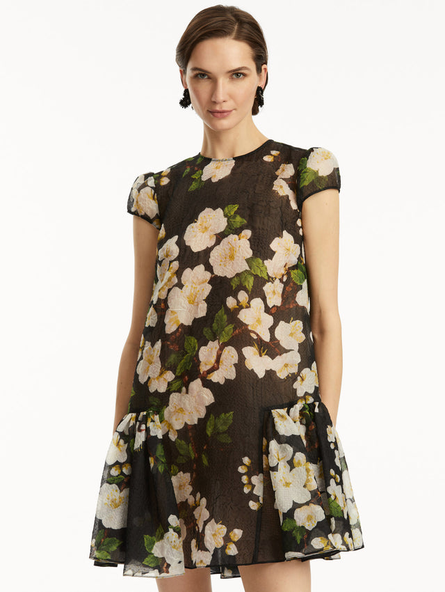 The model is wearing a short sleeve mini drop-waist dress in sheer black with white cherry blossoms printed all over. Zoom view.