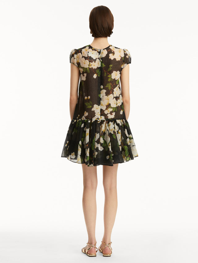 The model is wearing a short sleeve mini drop-waist dress in sheer black with white cherry blossoms printed all over. Back view.