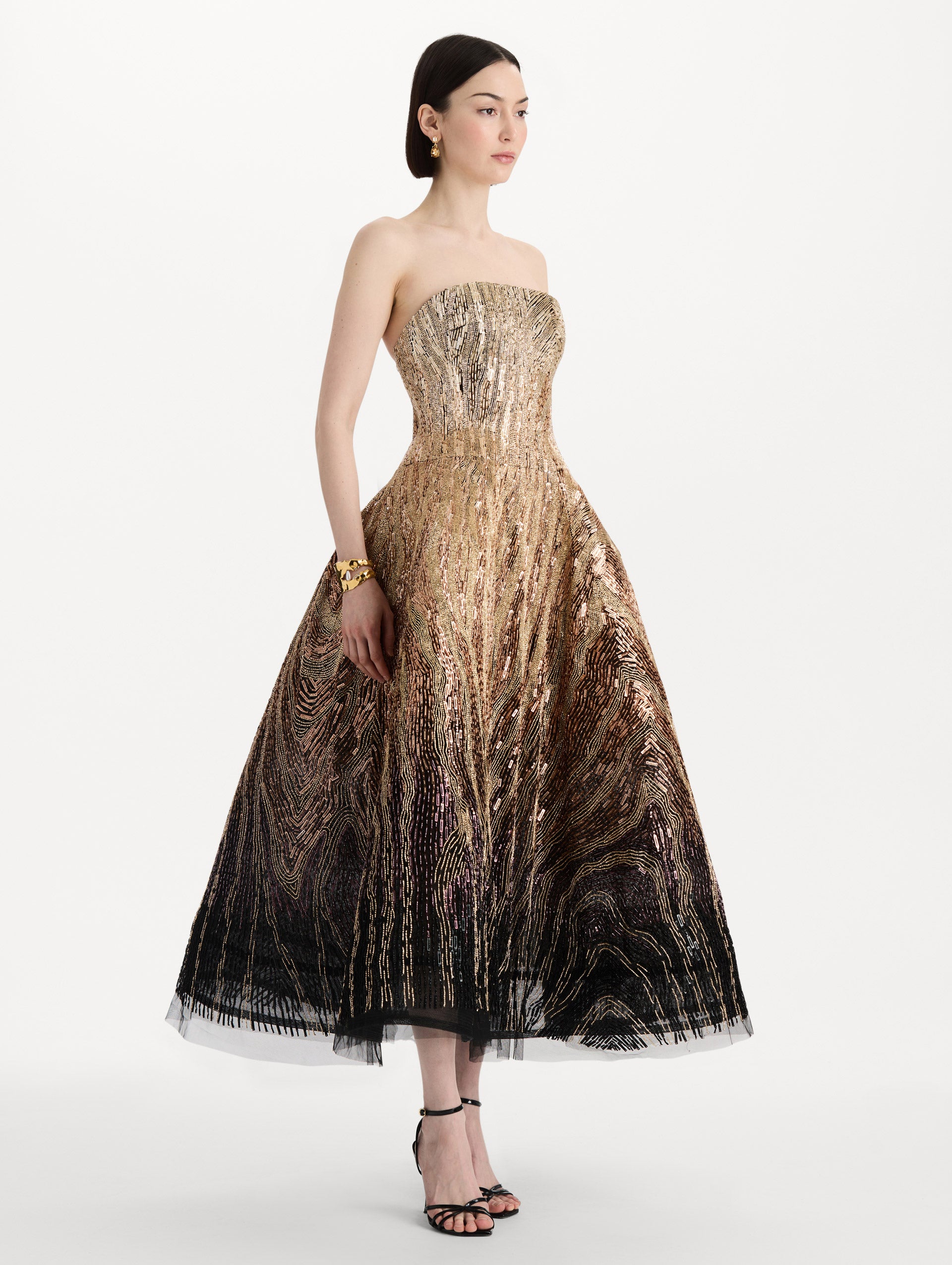 The model is wearing the beaded sequin moiré ombré tea length gown with a strapless silhouette and transitioning gold multi tones. Side view.