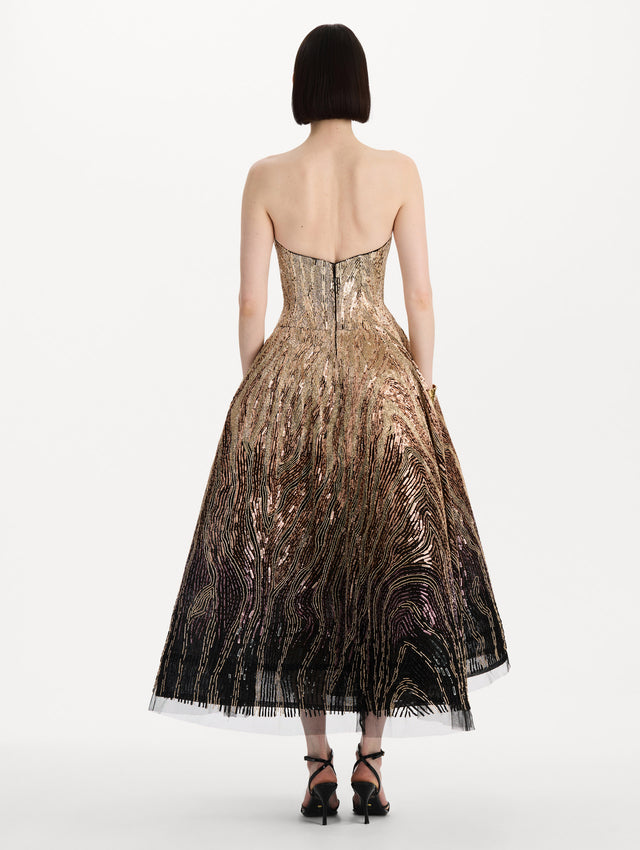 The model is wearing the beaded sequin moiré ombré tea length gown with a strapless silhouette and transitioning gold multi tones. Back view.