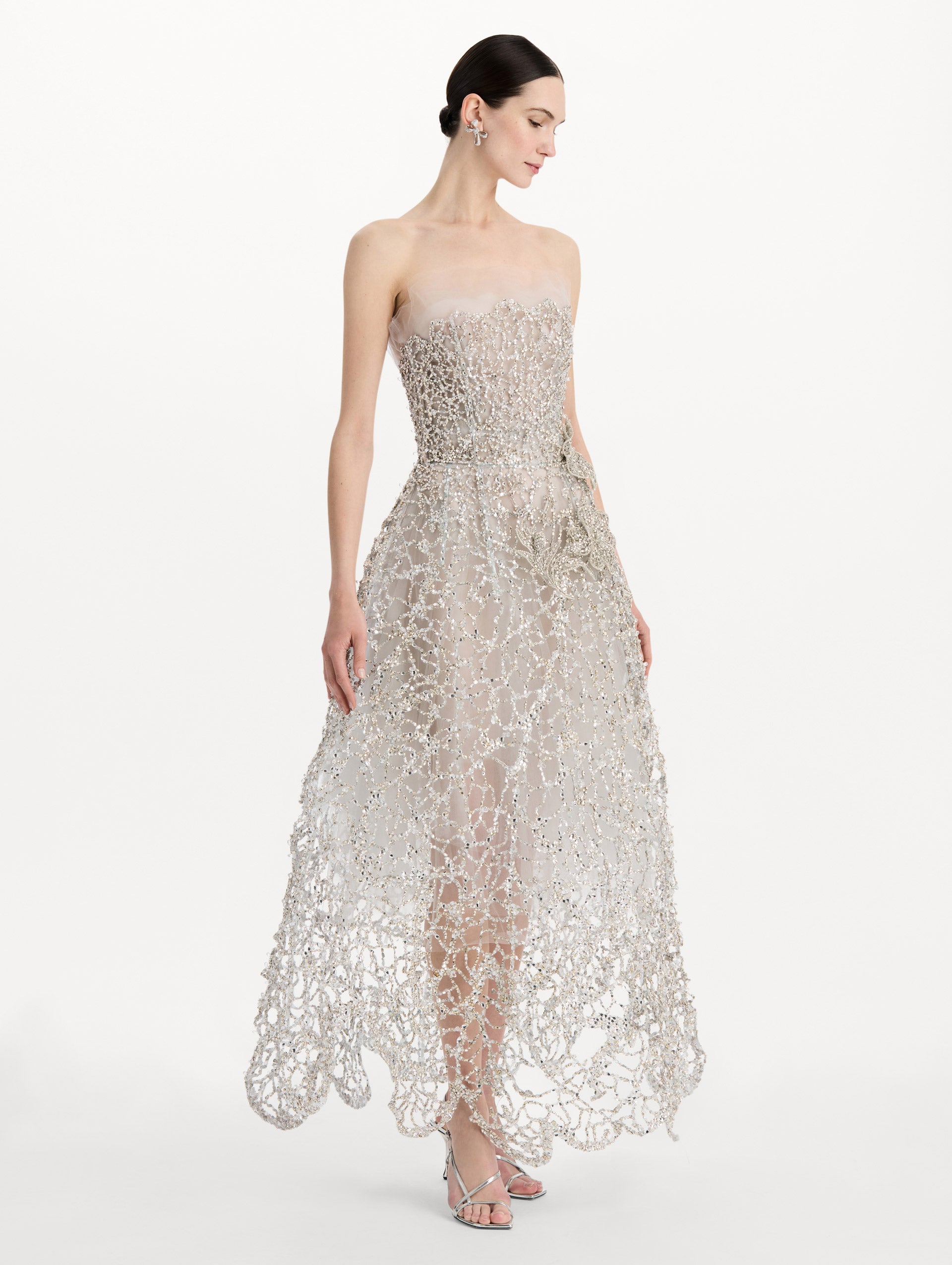The model is wearing the crystal leaves embroidered gown with a strapless silhouette and crystal leaf detailing in the color silver. Side view.