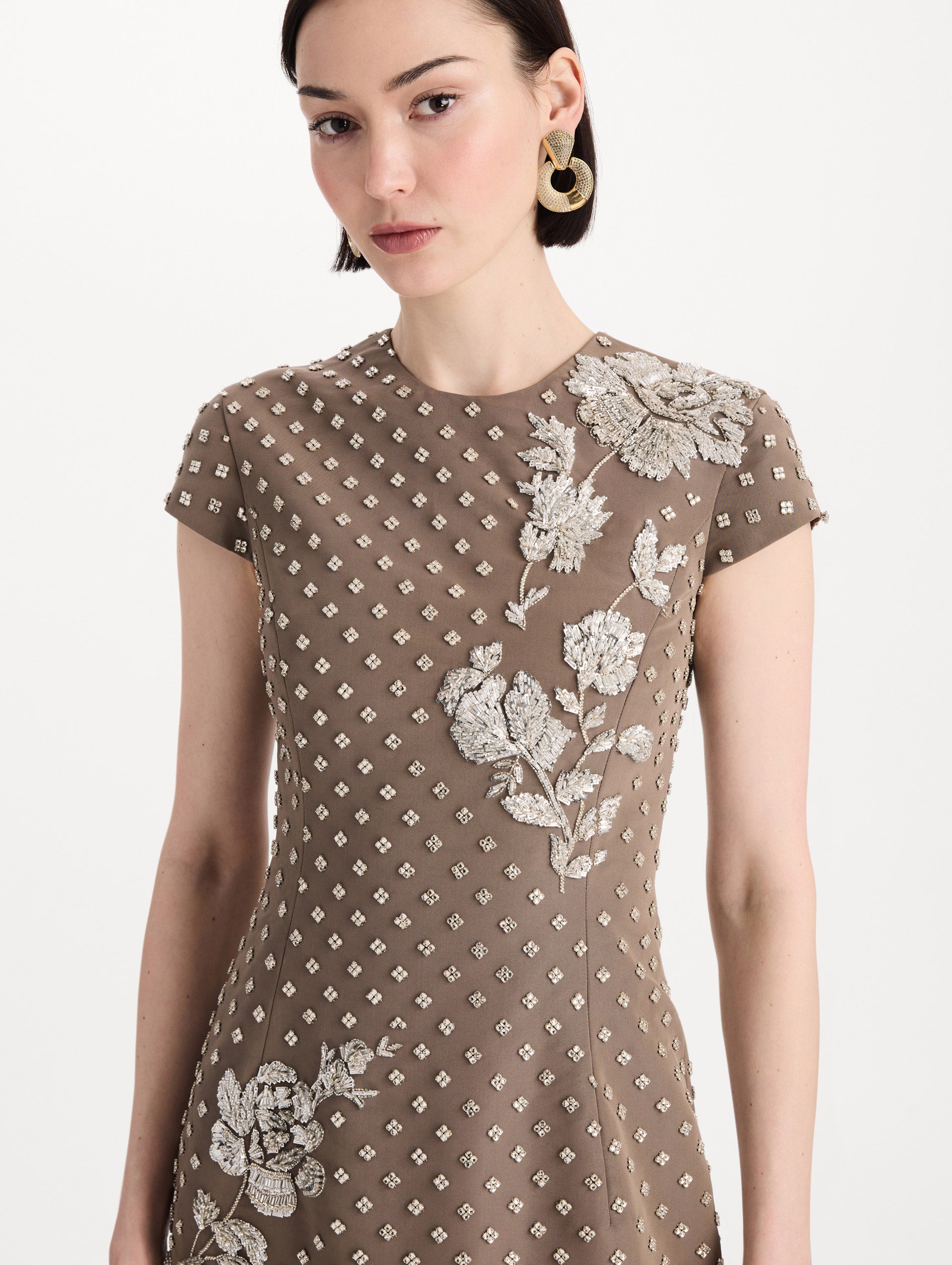 The model is wearing the crystal grid floral embroidered gown with short sleeves in the color stone. Zoom view. 