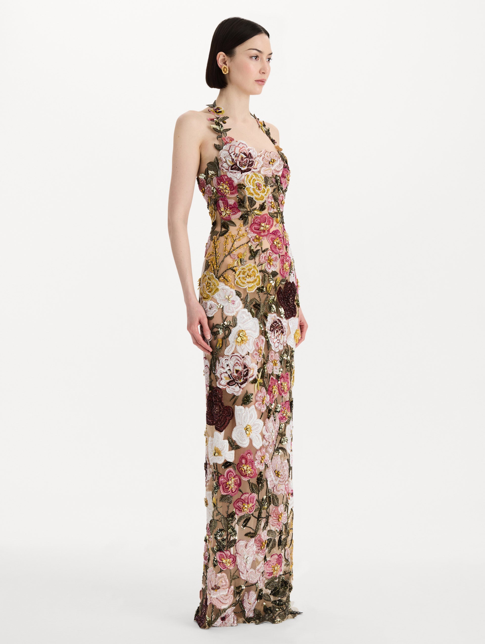 The model is wearing the beaded mixed botanical embroidered gown in multi-color with a halter neckline and floral detailing. Side view.