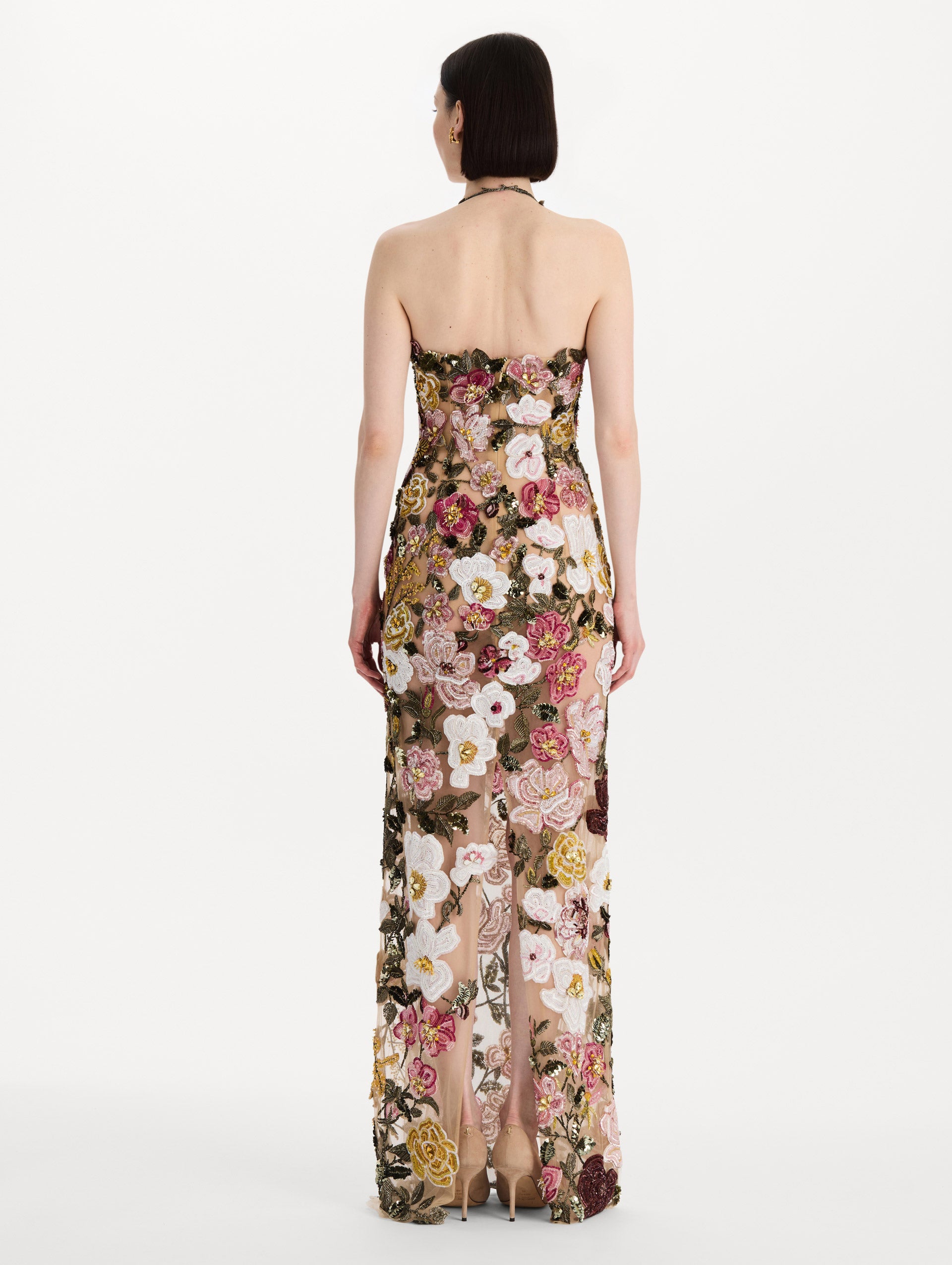The model is wearing the beaded mixed botanical embroidered gown in multi-color with a halter neckline and floral detailing. Back view.
