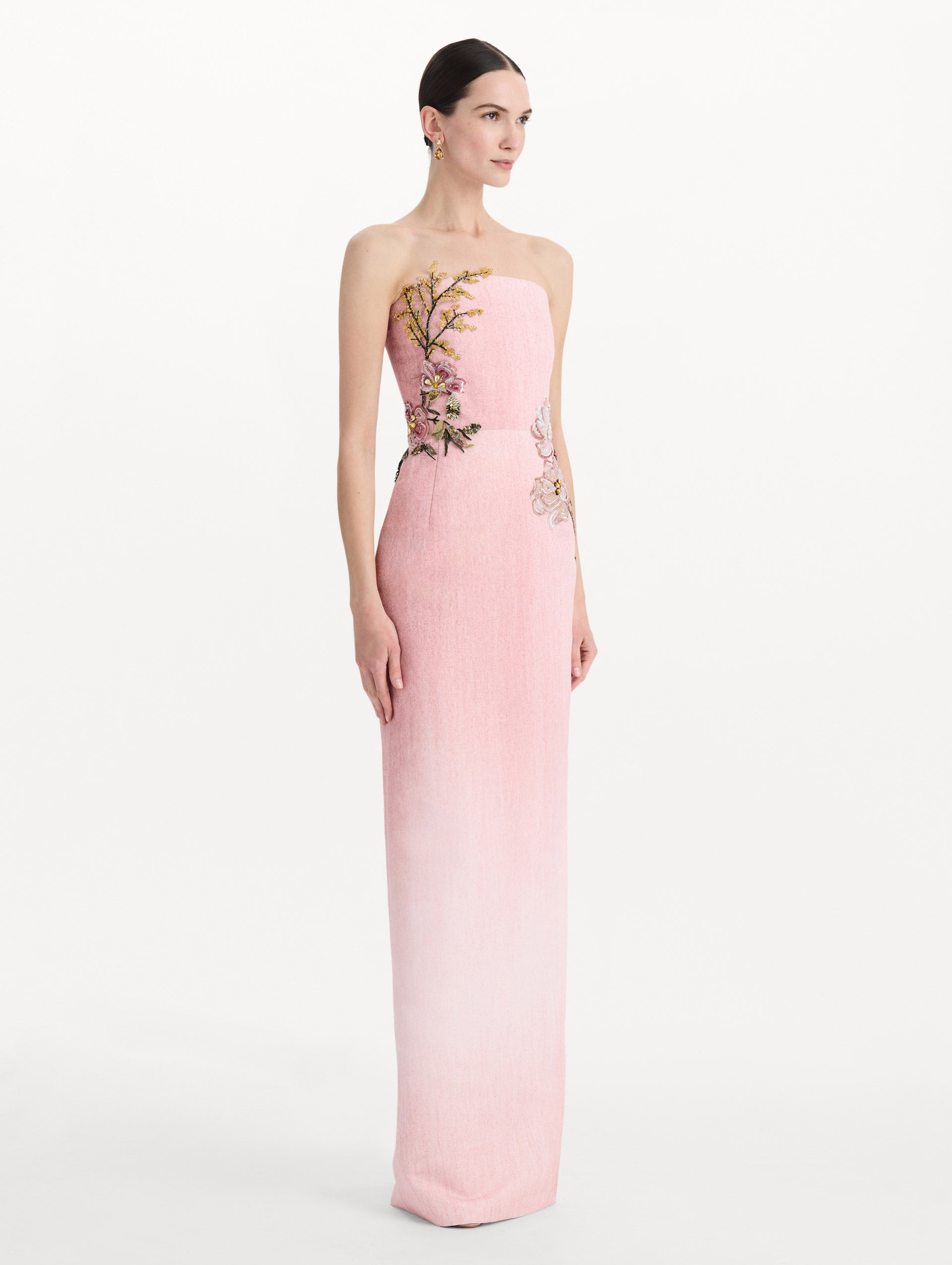 The model is wearing the beaded mixed botanical embroidered column gown with floral detailing and a strapless silhouette in the color blush. Side view.