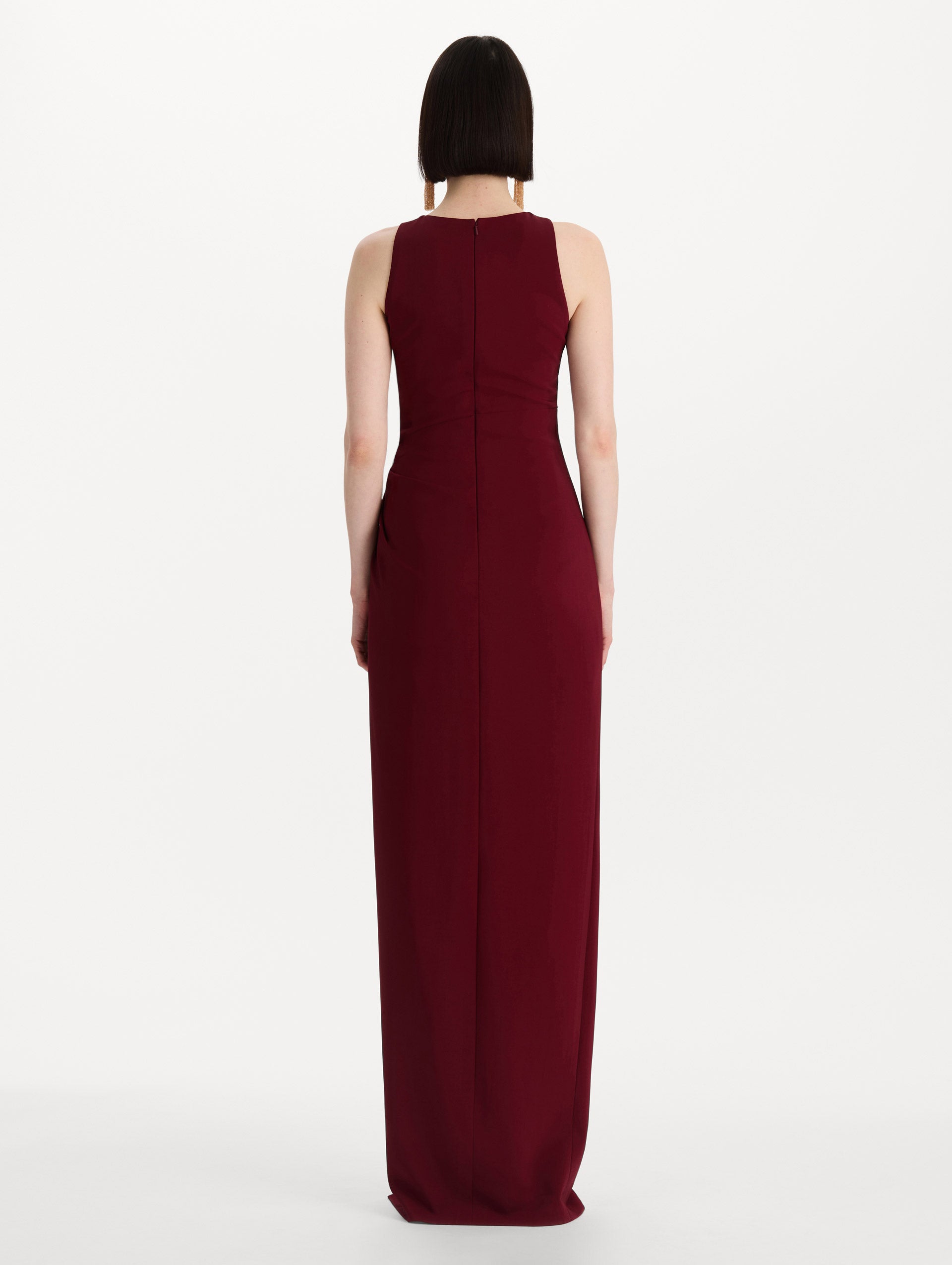 The model is wearing the drape-detailed cady gown with a sleeveless silhouette in the color claret. Back view. 