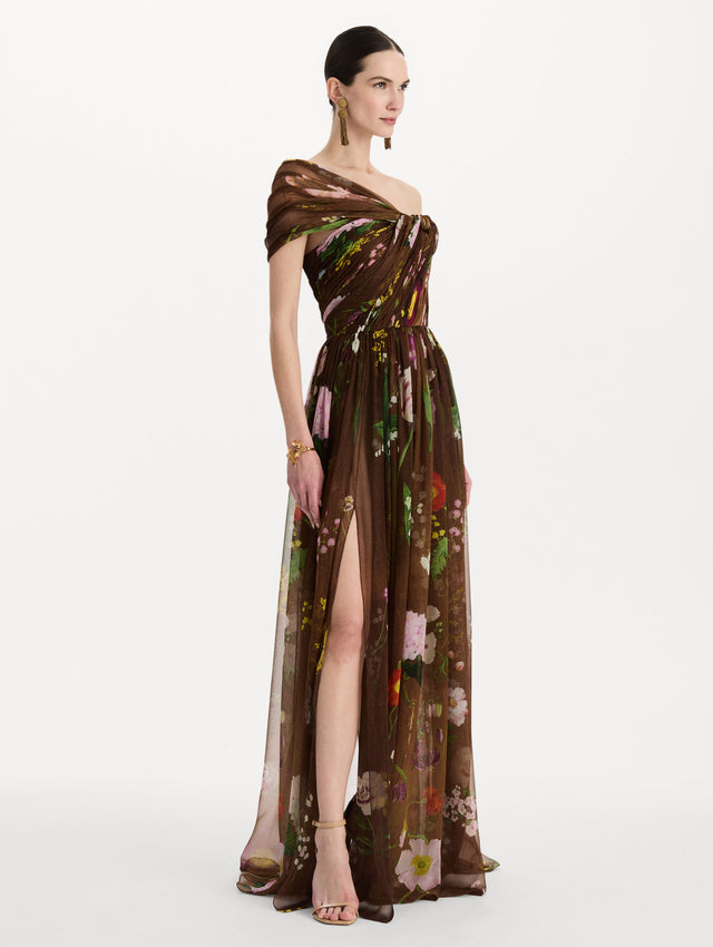 The model is wearing the mixed botanical silk chiffon gown with a one-shoulder neckline and a painterly floral print in a brown multi palette. Side view. 