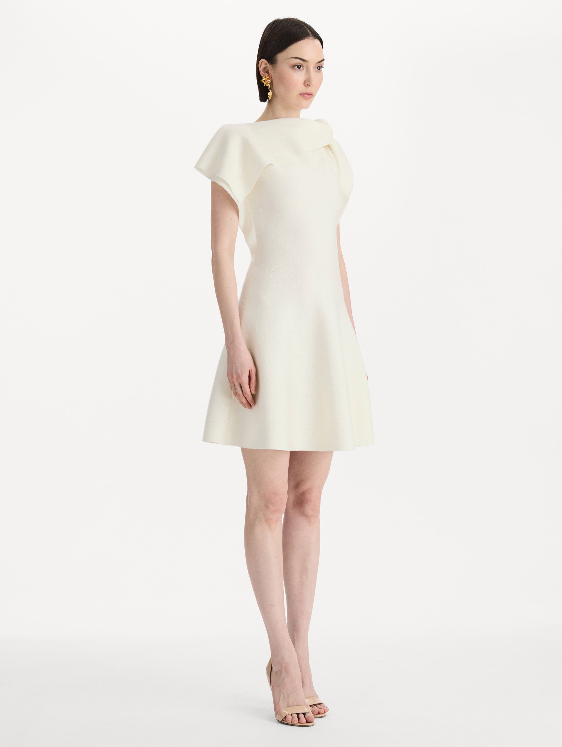 The model is wearing the drape-detailed knit dress with a draped neckline in the color ivory. Side view. 