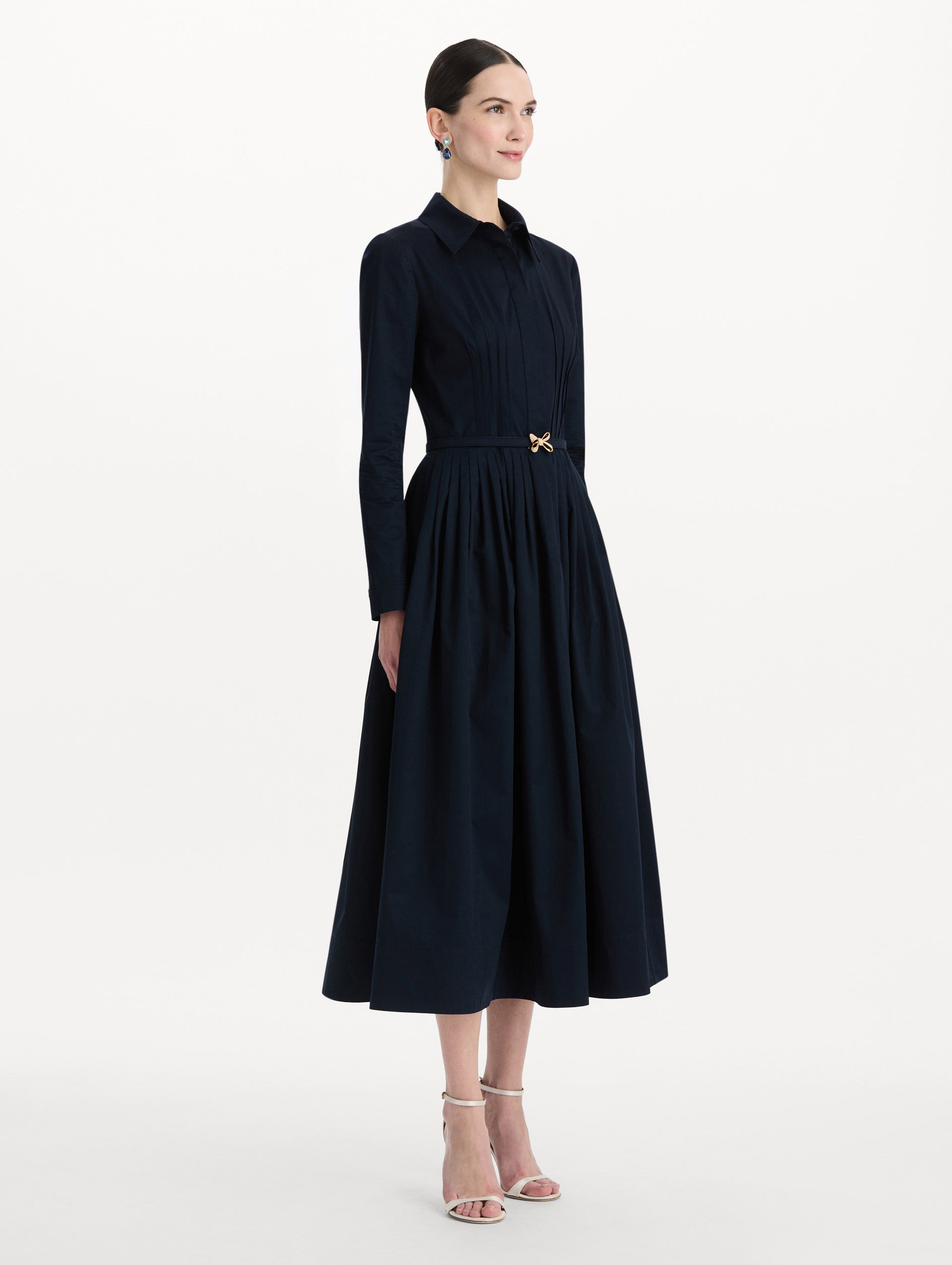 The model is wearing the bow belt cotton stretch twill dress with a collared neckline and long sleeves in the color navy. Side view. 