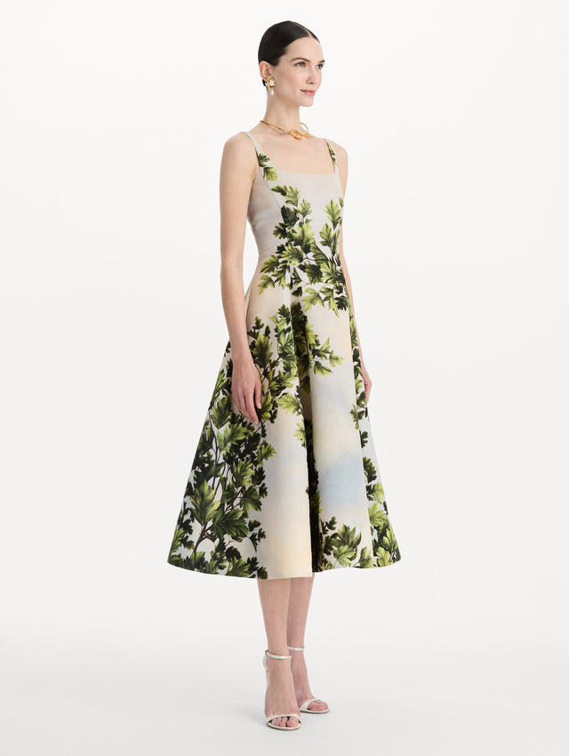 The model is wearing the maple leaves faille dress with a sleeveless scoop neckline and a foliage print in the color green multi. Side view. 