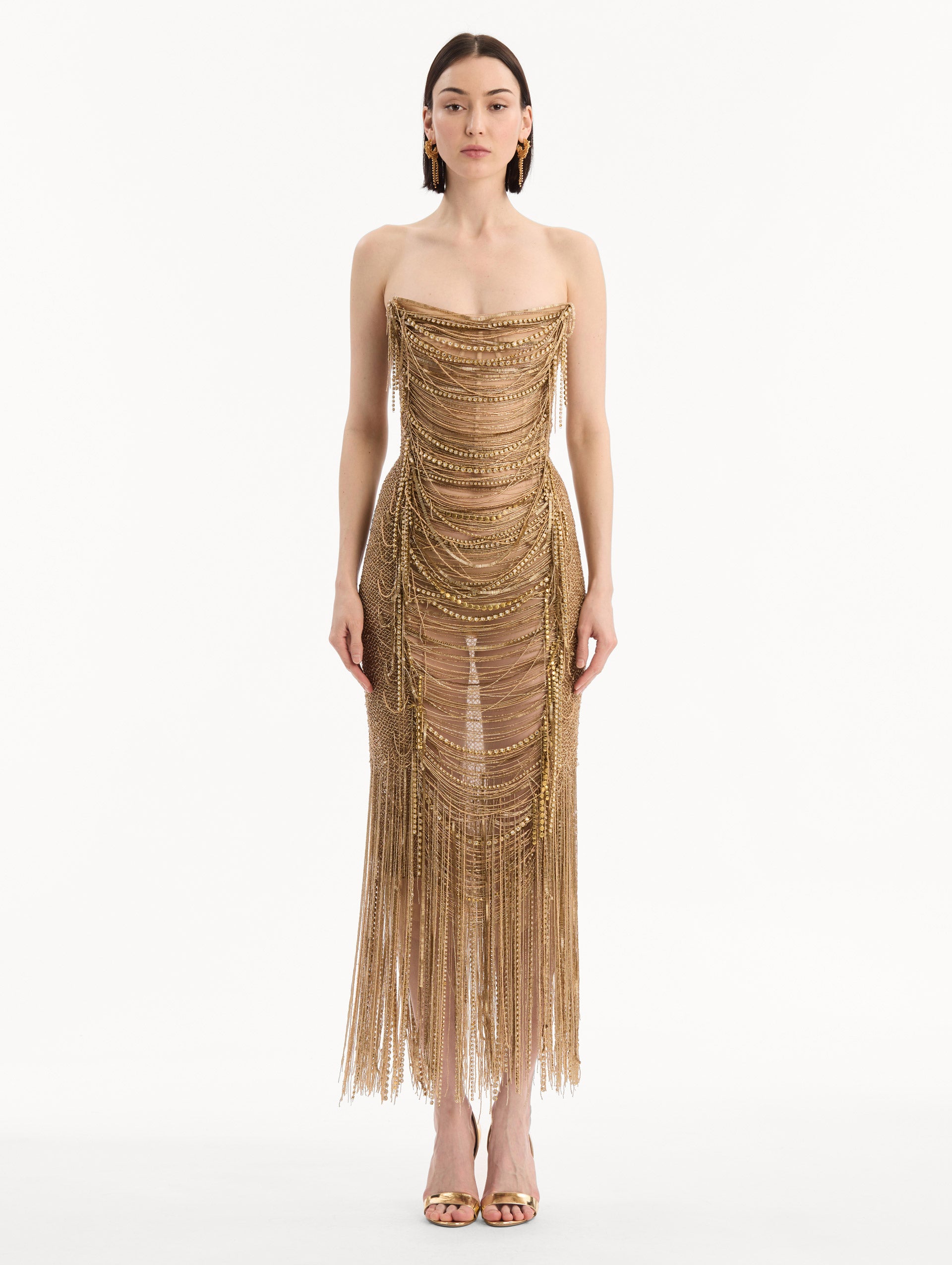 The model is wearing the Bugle Bead & Crystal Fringe Gown in Gold. Featuring a strapless neckline, layers of bugle beads and crystals draped across, and a fringe skirt. Front View.