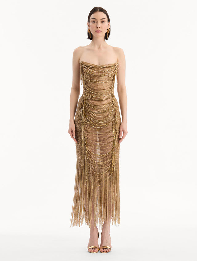 The model is wearing the Bugle Bead & Crystal Fringe Gown in Gold. Featuring a strapless neckline, layers of bugle beads and crystals draped across, and a fringe skirt. Front View.