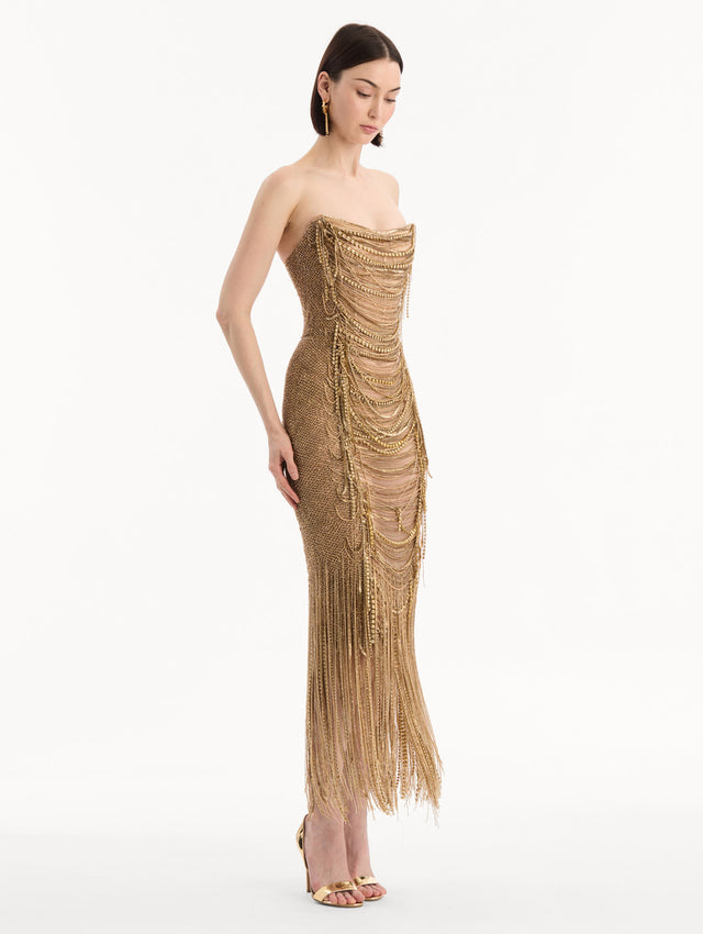 The model is wearing the Bugle Bead & Crystal Fringe Gown in Gold. Featuring a strapless neckline, layers of bugle beads and crystals draped across, and a fringe skirt. Side View.
