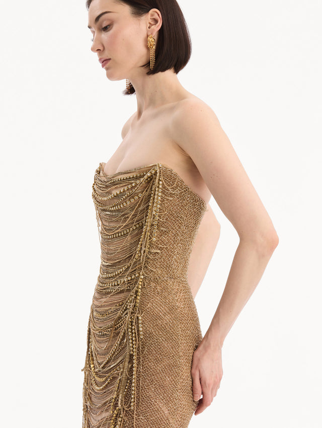 The model is wearing the Bugle Bead & Crystal Fringe Gown in Gold. Featuring a strapless neckline, layers of bugle beads and crystals draped across, and a fringe skirt. Detail View.