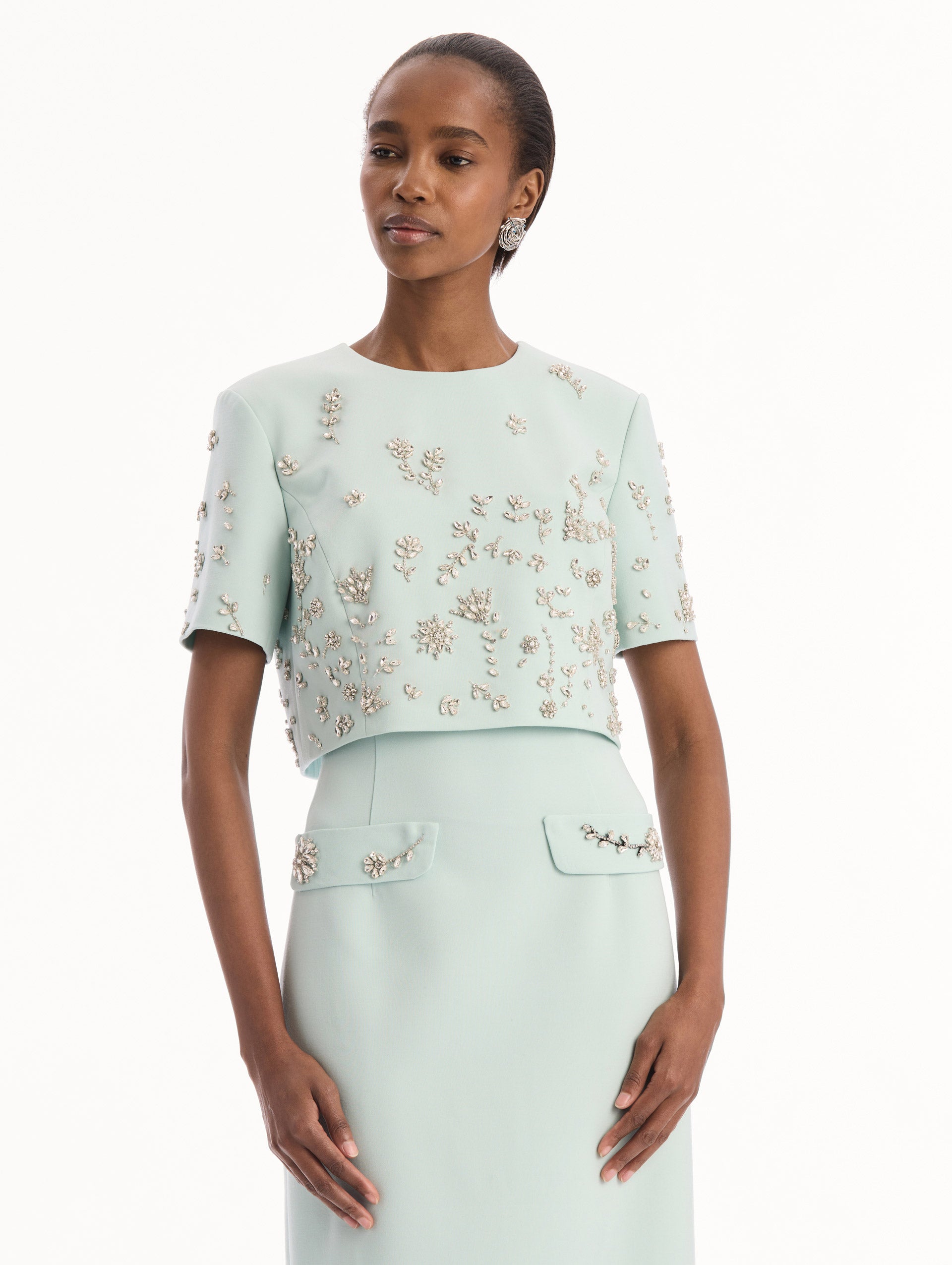 The model is wearing the Crystal Floral Embroidered Gown in Saltwater Blue. Featuring a strapless neckline, faux front flap pockets with crystal embroidery, and a long, column silhouette. Detail View.