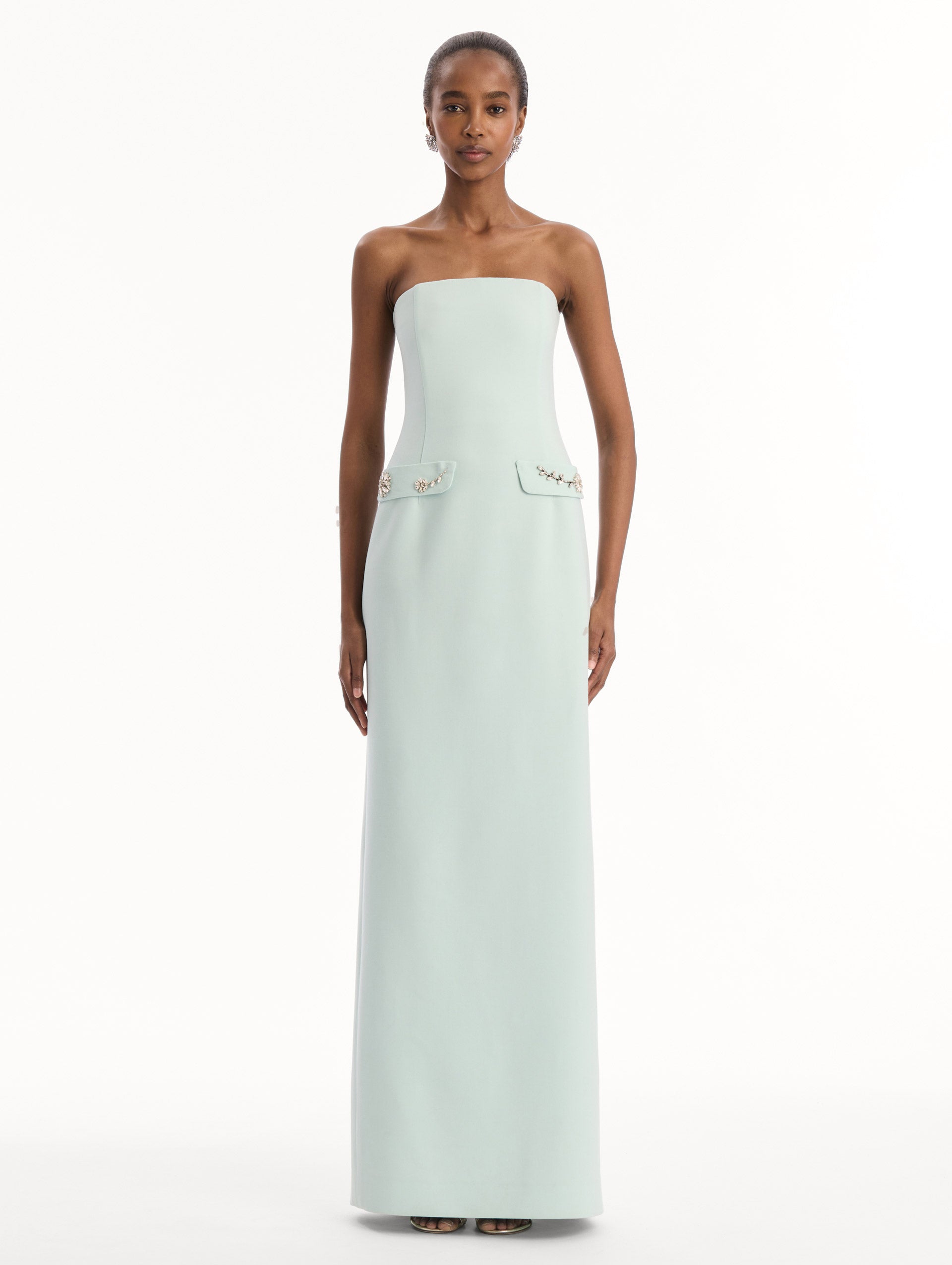 The model is wearing the Crystal Floral Embroidered Gown in Saltwater Blue. Featuring a strapless neckline, faux front flap pockets with crystal embroidery, and a long, column silhouette. Front View.