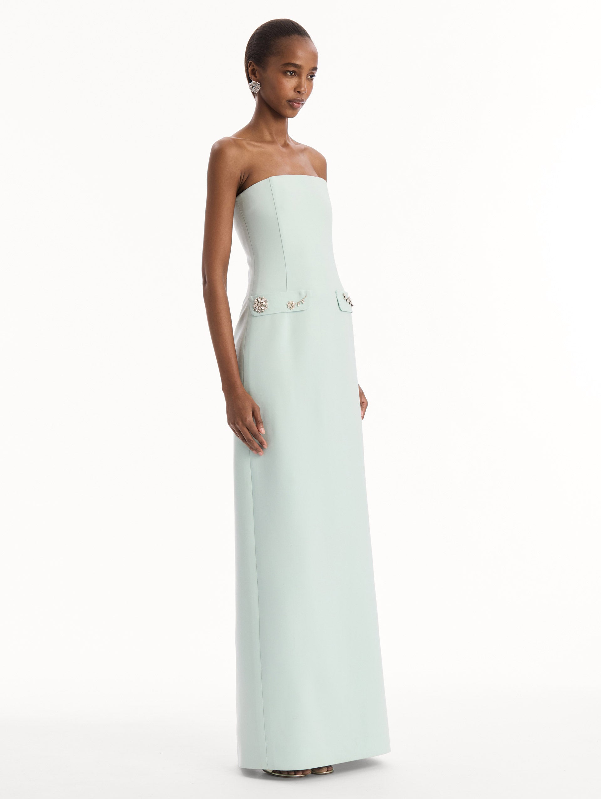 The model is wearing the Crystal Floral Embroidered Gown in Saltwater Blue. Featuring a strapless neckline, faux front flap pockets with crystal embroidery, and a long, column silhouette. Side View.