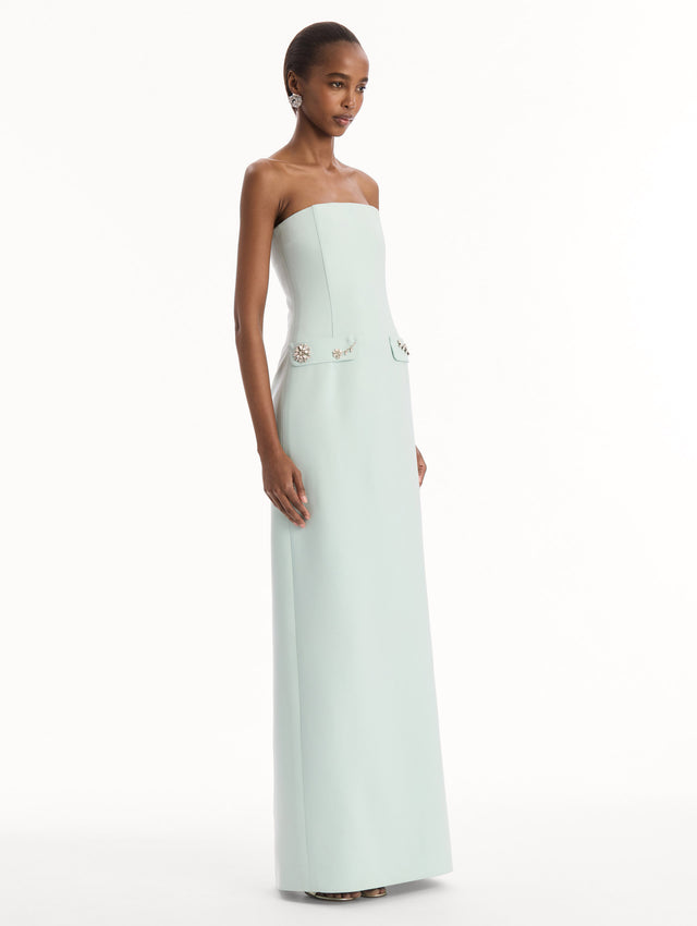 The model is wearing the Crystal Floral Embroidered Gown in Saltwater Blue. Featuring a strapless neckline, faux front flap pockets with crystal embroidery, and a long, column silhouette. Side View.