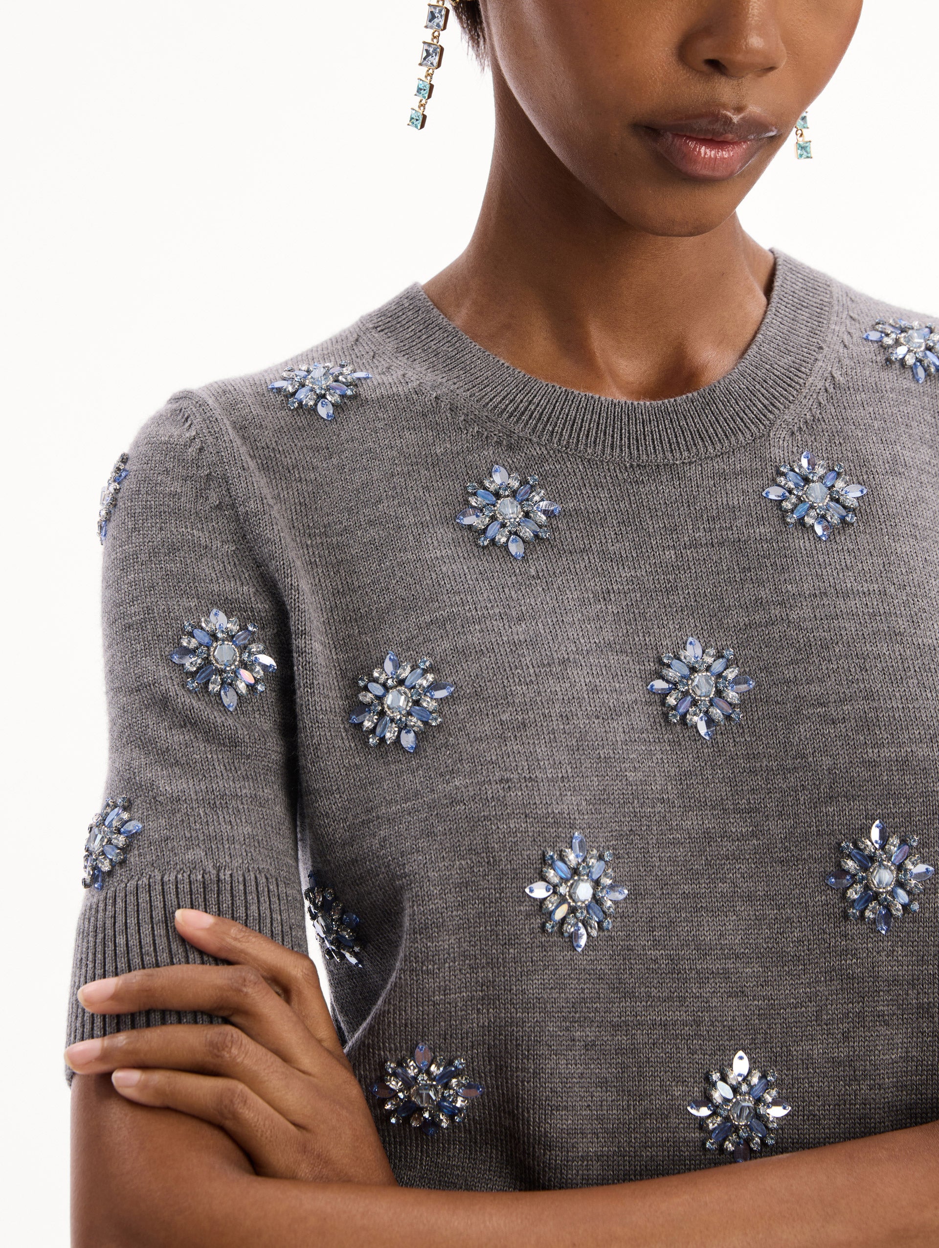 The model is wearing the Crystal Embroidered Pullover in grey with blue crystal embroidered. Featuring a crew neckline and ribbed trims. Detail View.