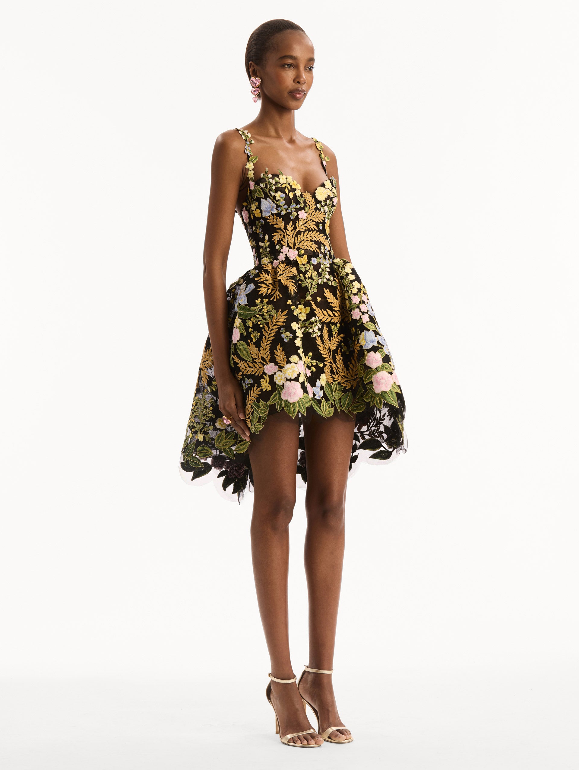 The model is wearing the Dominican Mixed-Floral Guipure High-Low Dress. Featuring a sweetheart neckline, delicate floral straps, fitted bodice, and mini skirt. Side View.