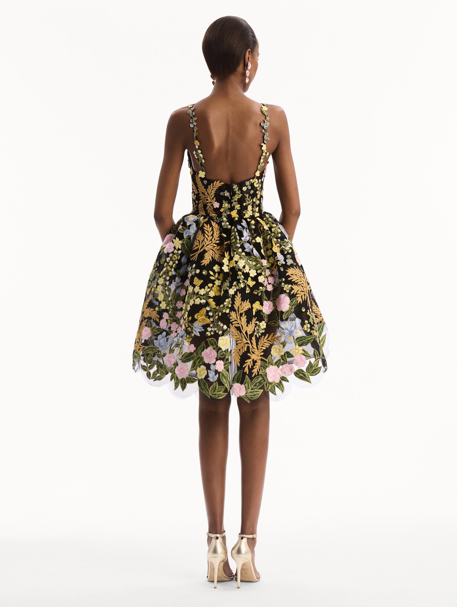 The model is wearing the Dominican Mixed-Floral Guipure High-Low Dress. Featuring a sweetheart neckline, delicate floral straps, fitted bodice, and mini skirt. Back View.