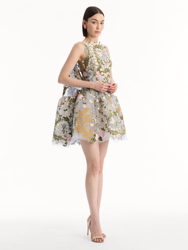The model is wearing the Dominican Mixed-Floral Guipure Cape-Back Dress. Featuring a straight neckline, fitted bodice, flared mini skirt, and structural cape. Side View.