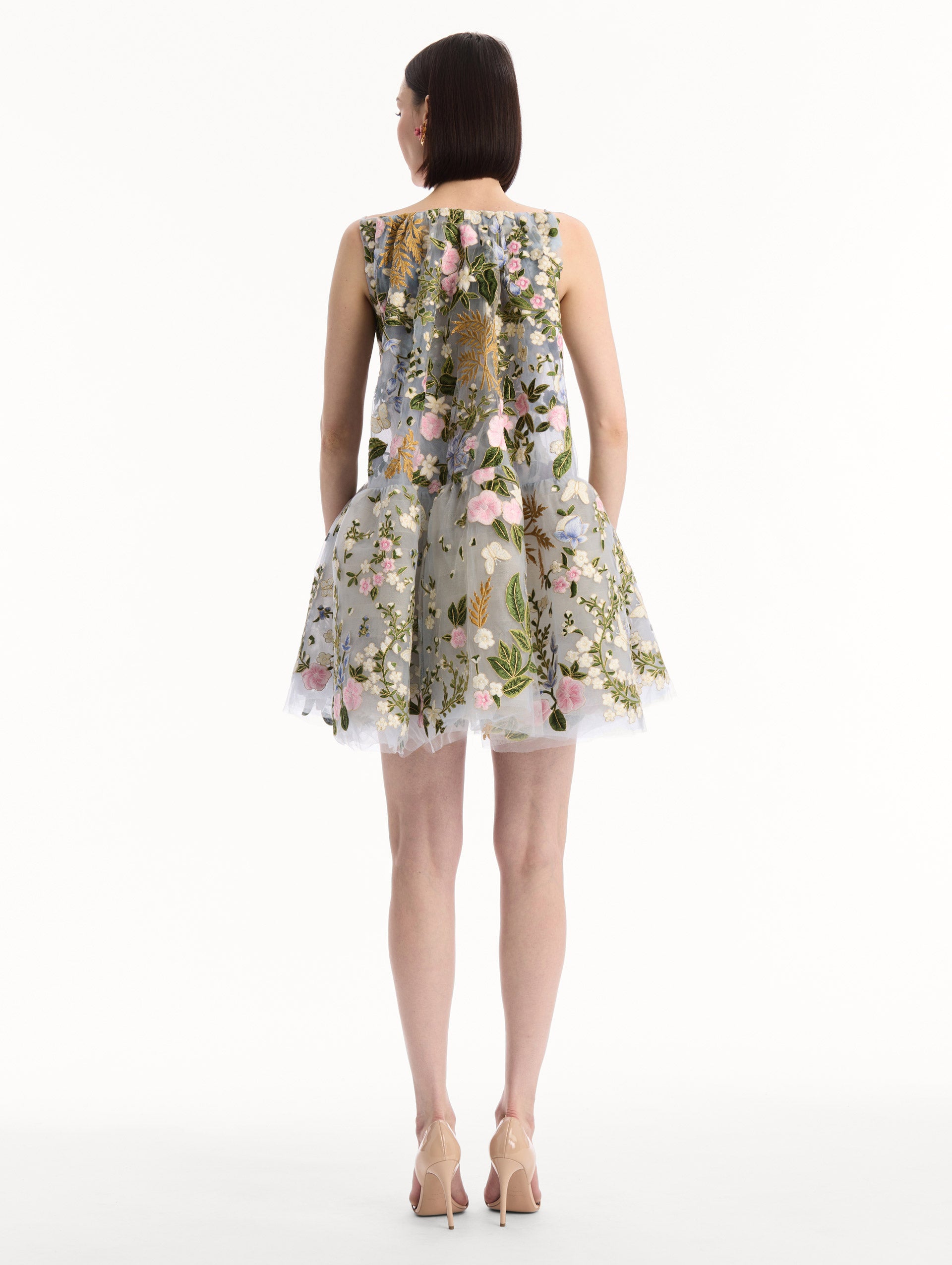 The model is wearing the Dominican Mixed-Floral Guipure Cape-Back Dress. Featuring a straight neckline, fitted bodice, flared mini skirt, and structural cape. Back View.