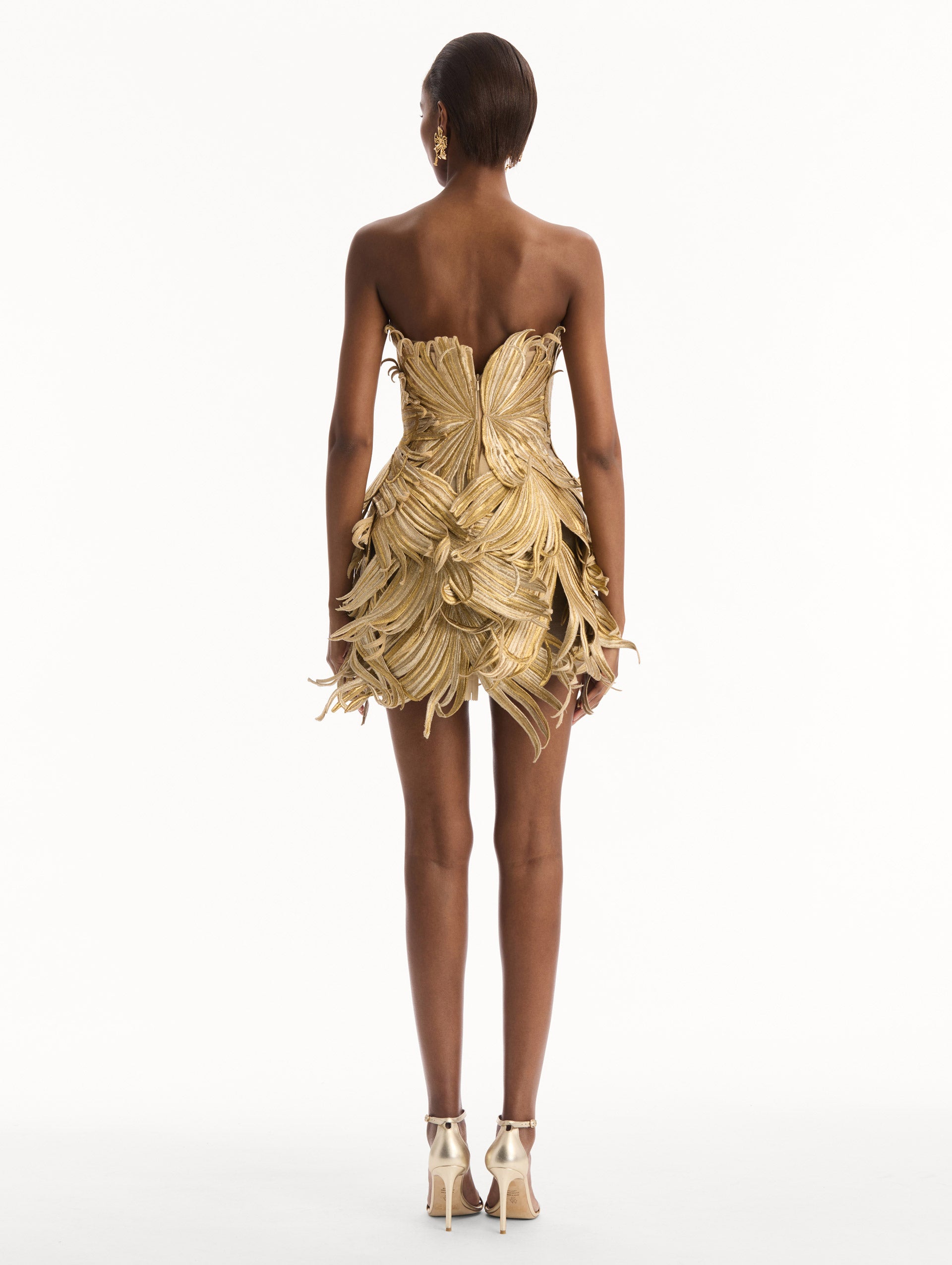The model is wearing the Palm Leaves Metallic Cocktail Dress in Gold. Featuring dynamic palm leaves sculpted in metallic embroidery and a mini-length skirt. Back View.