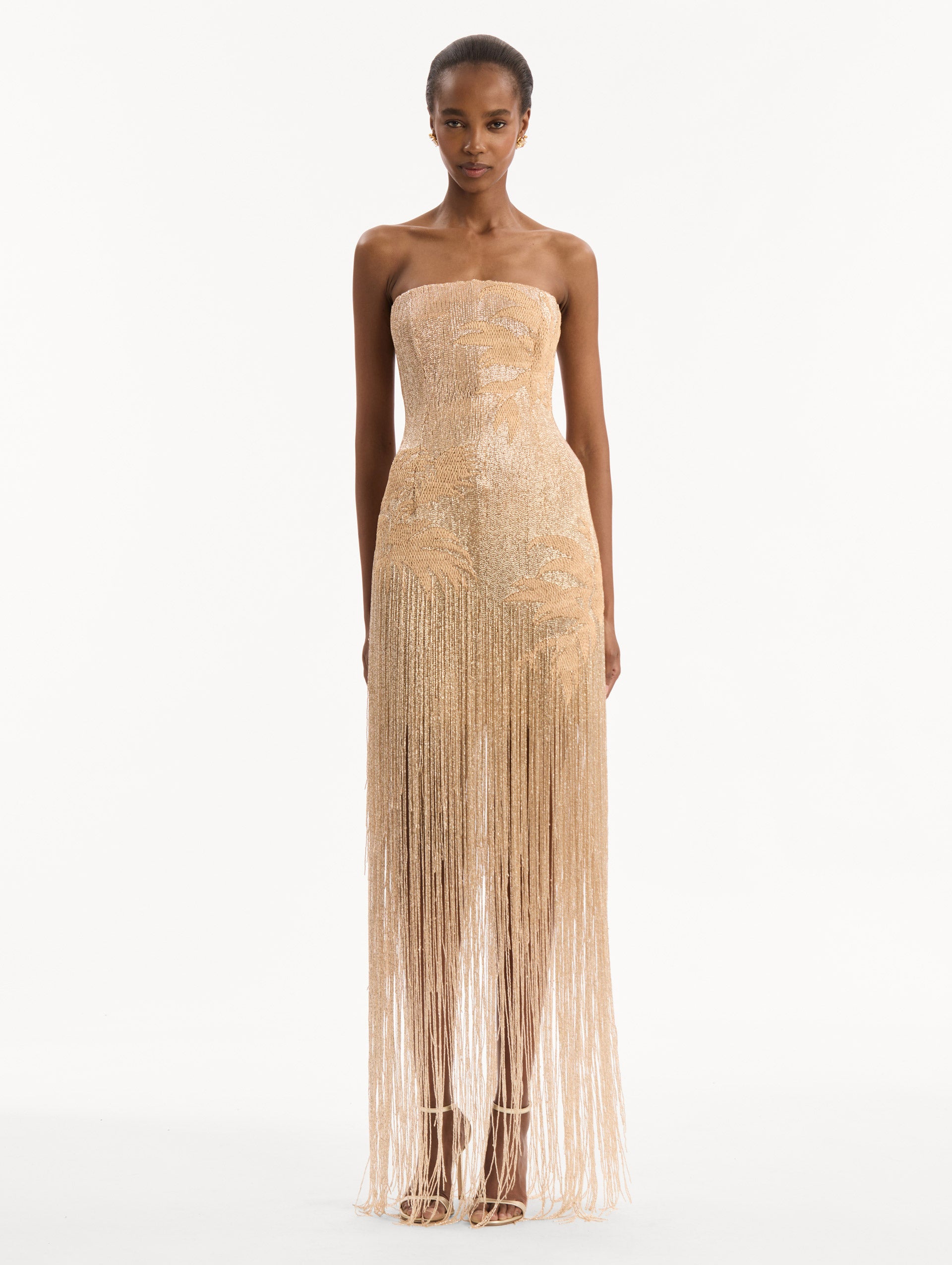 The model is wearing the Palm Tree Raffia Fringe Gown. Featuring a strapless neckline, raffia embroidery, and long, column silhouette with fringe skirt. Front View.