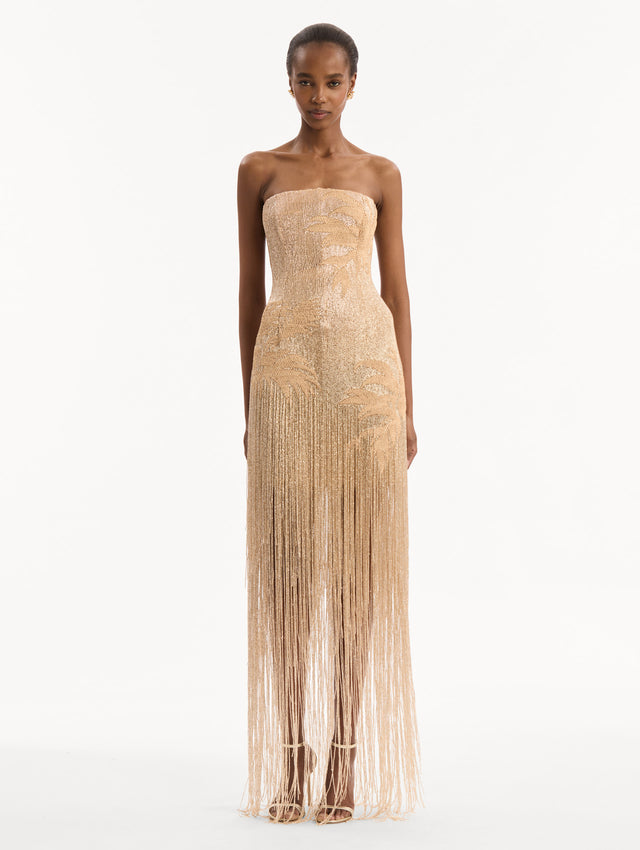 The model is wearing the Palm Tree Raffia Fringe Gown. Featuring a strapless neckline, raffia embroidery, and long, column silhouette with fringe skirt. Front View.