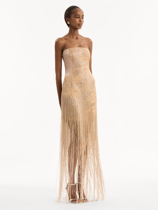 The model is wearing the Palm Tree Raffia Fringe Gown. Featuring a strapless neckline, raffia embroidery, and long, column silhouette with fringe skirt. Side View.