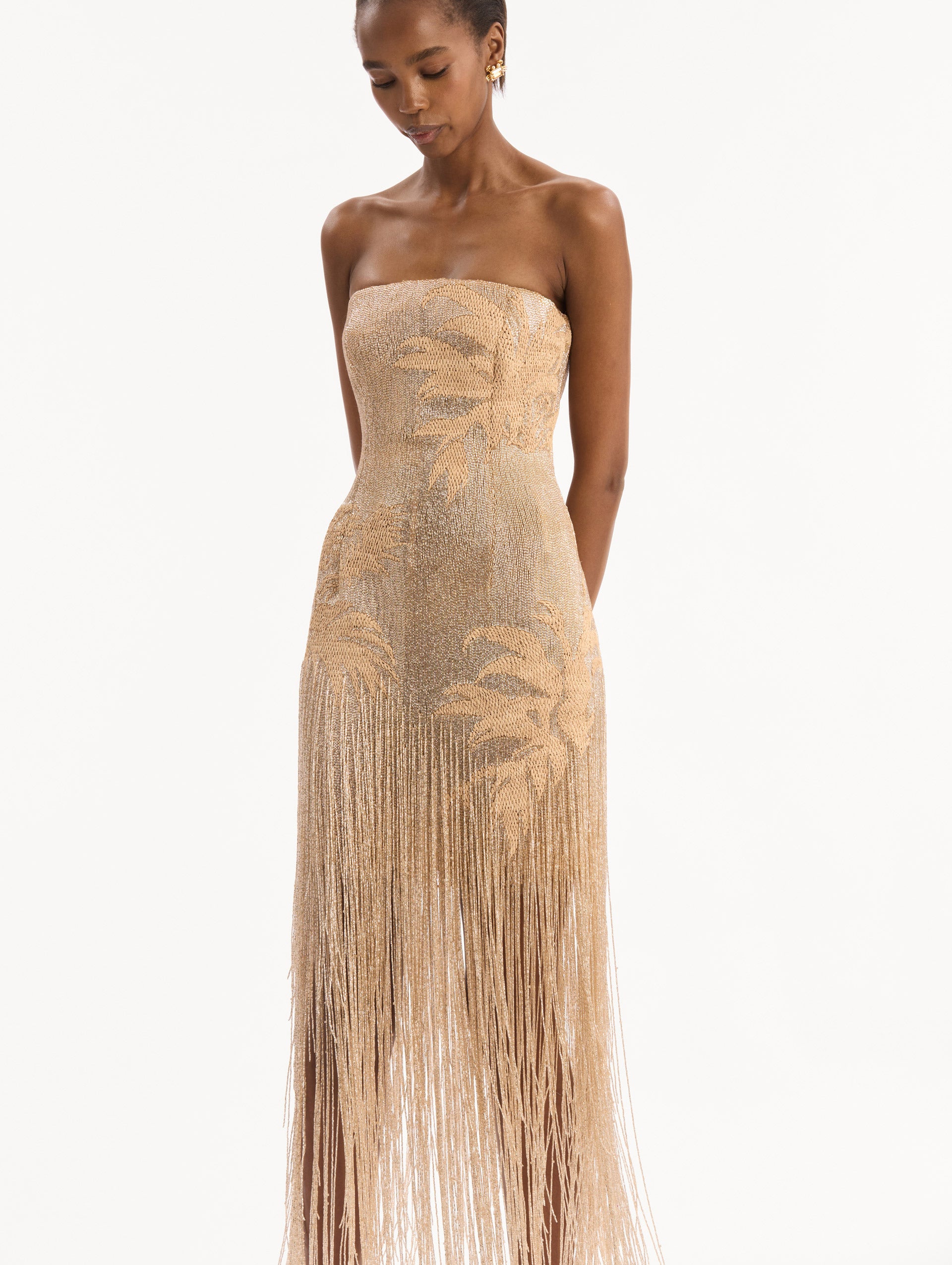 The model is wearing the Palm Tree Raffia Fringe Gown. Featuring a strapless neckline, raffia embroidery, and long, column silhouette with fringe skirt. Detail View.