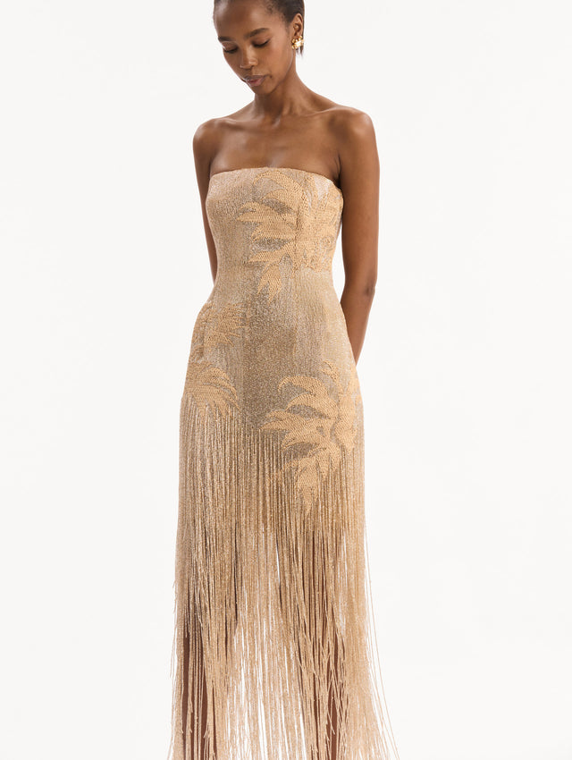 The model is wearing the Palm Tree Raffia Fringe Gown. Featuring a strapless neckline, raffia embroidery, and long, column silhouette with fringe skirt. Detail View.