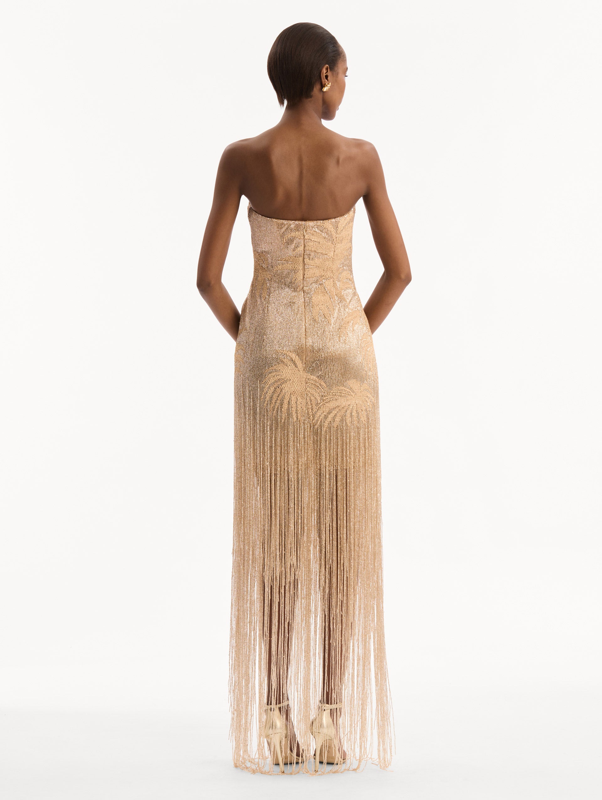 The model is wearing the Palm Tree Raffia Fringe Gown. Featuring a strapless neckline, raffia embroidery, and long, column silhouette with fringe skirt. Back View.