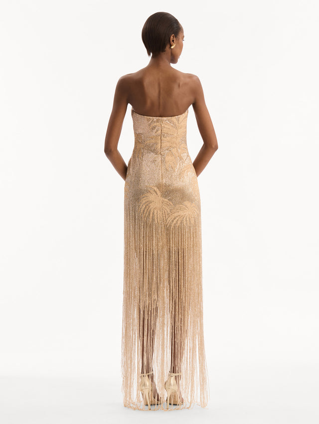 The model is wearing the Palm Tree Raffia Fringe Gown. Featuring a strapless neckline, raffia embroidery, and long, column silhouette with fringe skirt. Back View.
