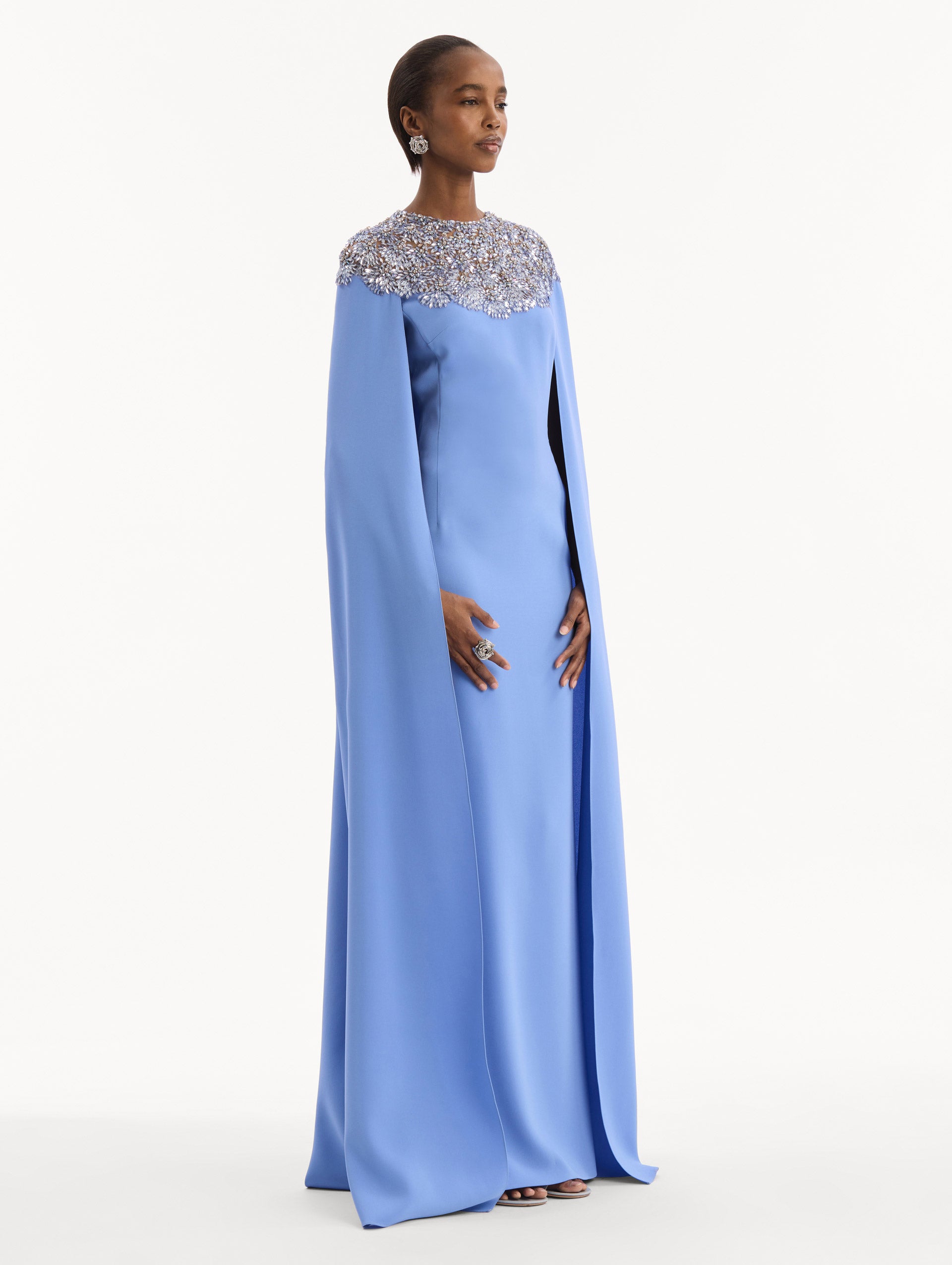 The model is wearing the Crystal & Glass Embroidered Caftan in a hydrangea blue color. Featuring crystal and glass bead embroidery at the collar, flowing cape-like sleeves, and a long skirt. Side View.