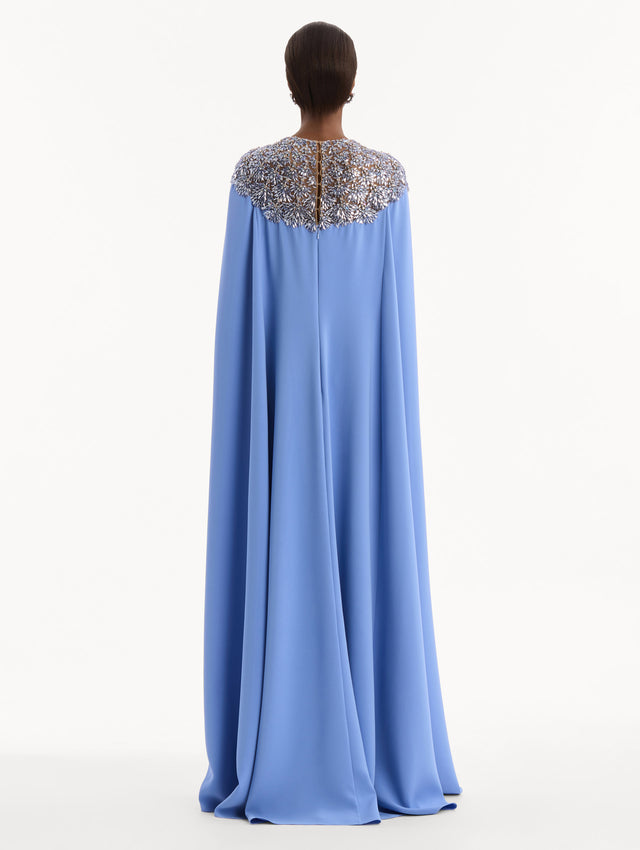 The model is wearing the Crystal & Glass Embroidered Caftan in a hydrangea blue color. Featuring crystal and glass bead embroidery at the collar, flowing cape-like sleeves, and a long skirt. Back View.