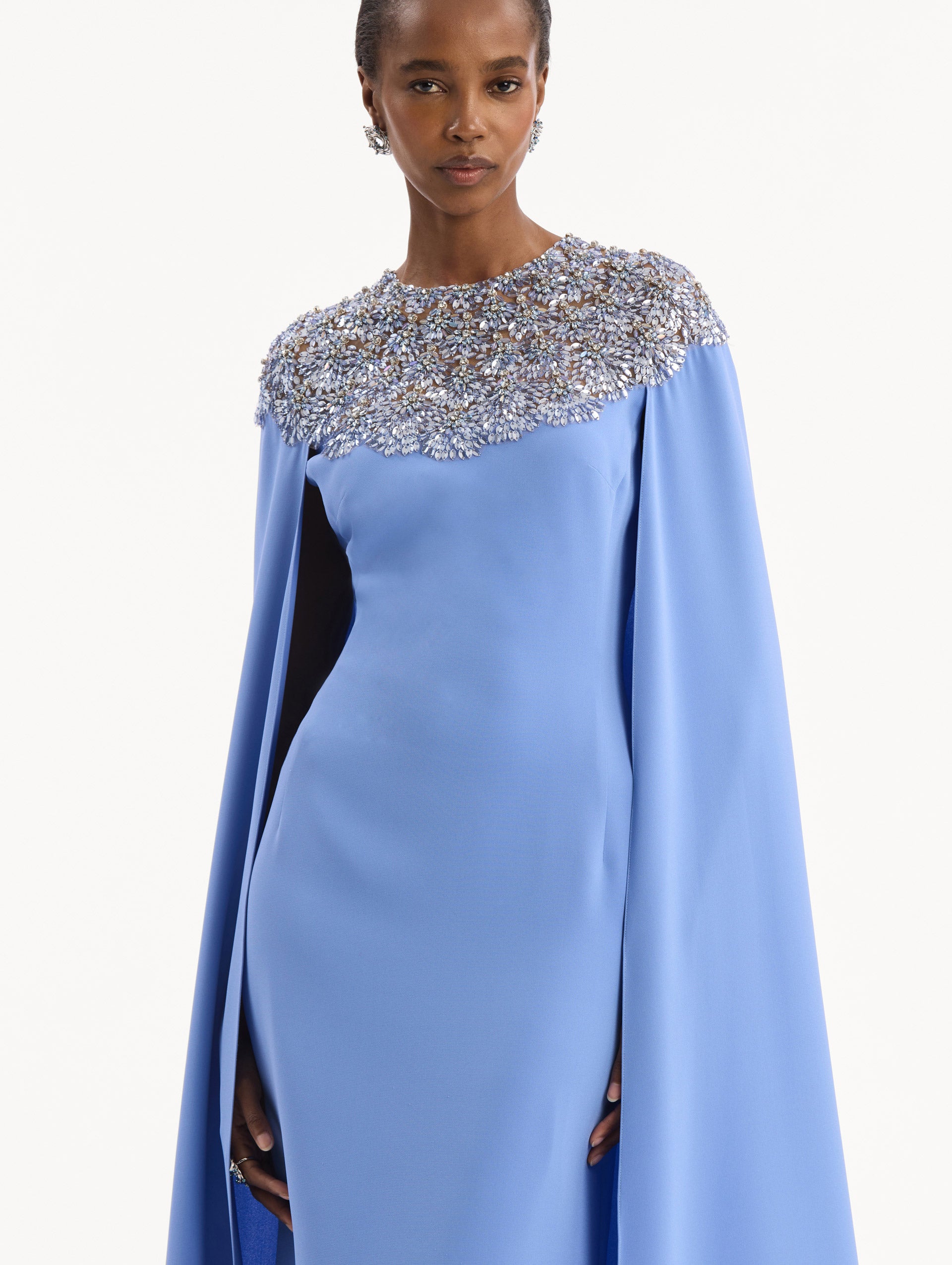 The model is wearing the Crystal & Glass Embroidered Caftan in a hydrangea blue color. Featuring crystal and glass bead embroidery at the collar, flowing cape-like sleeves, and a long skirt. Detail View.